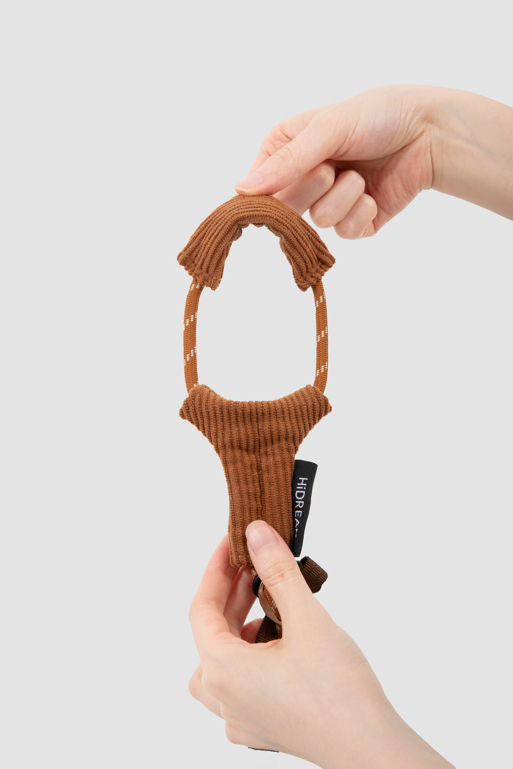 A person's hands holding a brown ribbed knit cat harness against a light gray background. The toy features a looped top and a textured body with a small black tag that reads 'HIDREAM'.