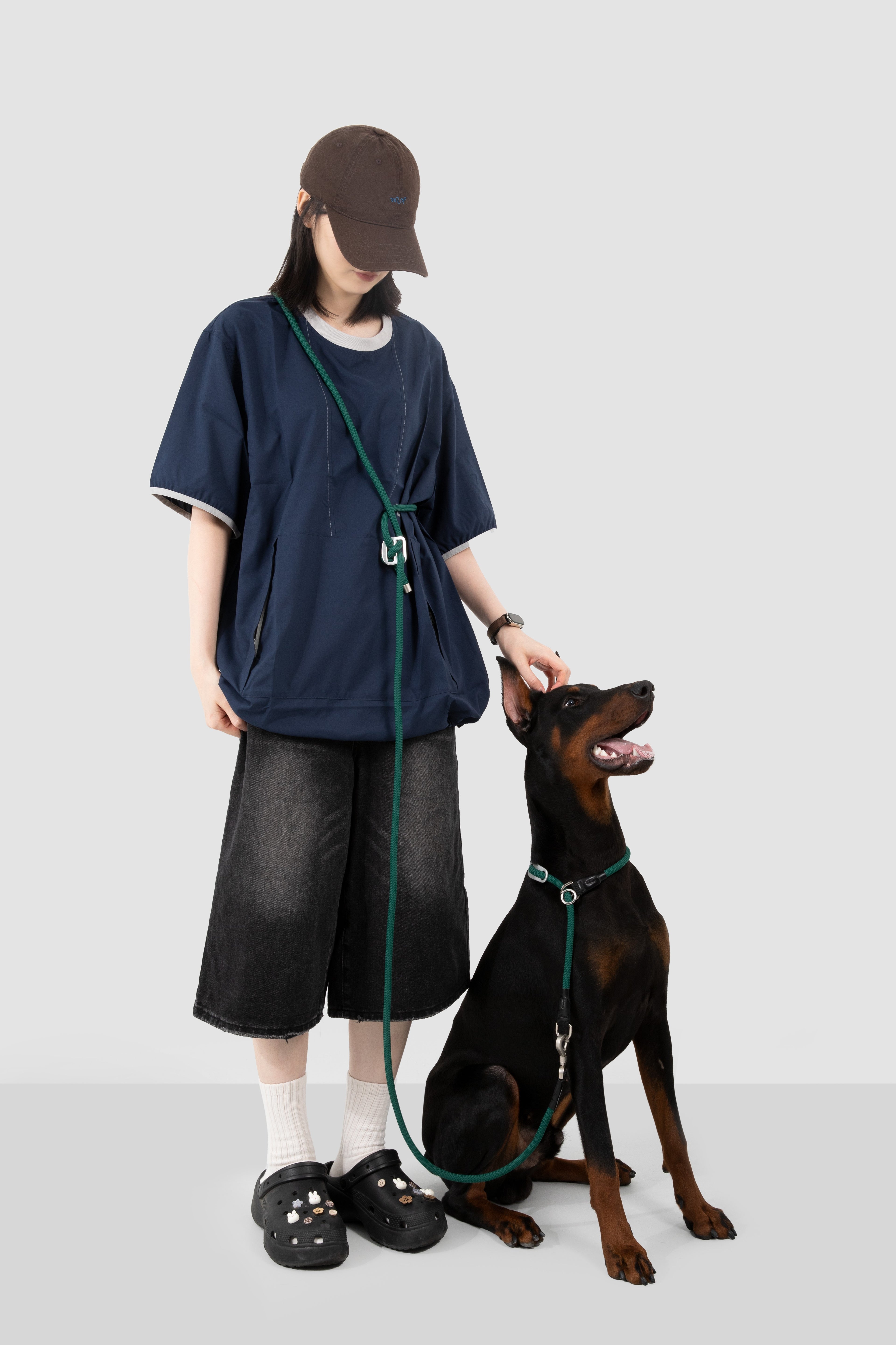 A person wearing a navy oversized T-shirt, dark denim shorts, white socks, and black slip-on shoes stands next to a black and brown dog on a green leash, against a light gray studio background.