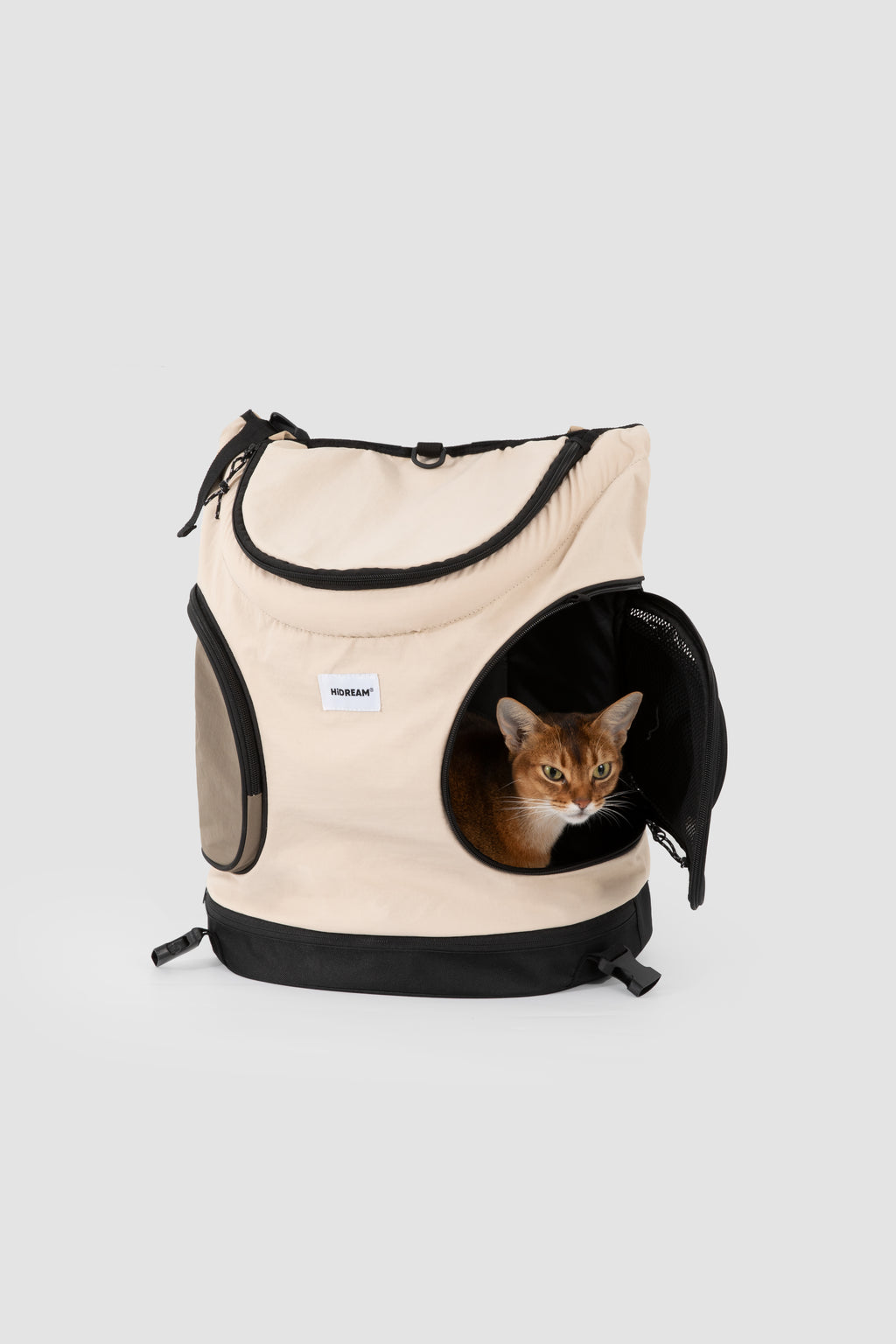 A beige and black pet backpack with a cat peeking out of its circular front opening, photographed on a light gray studio background.