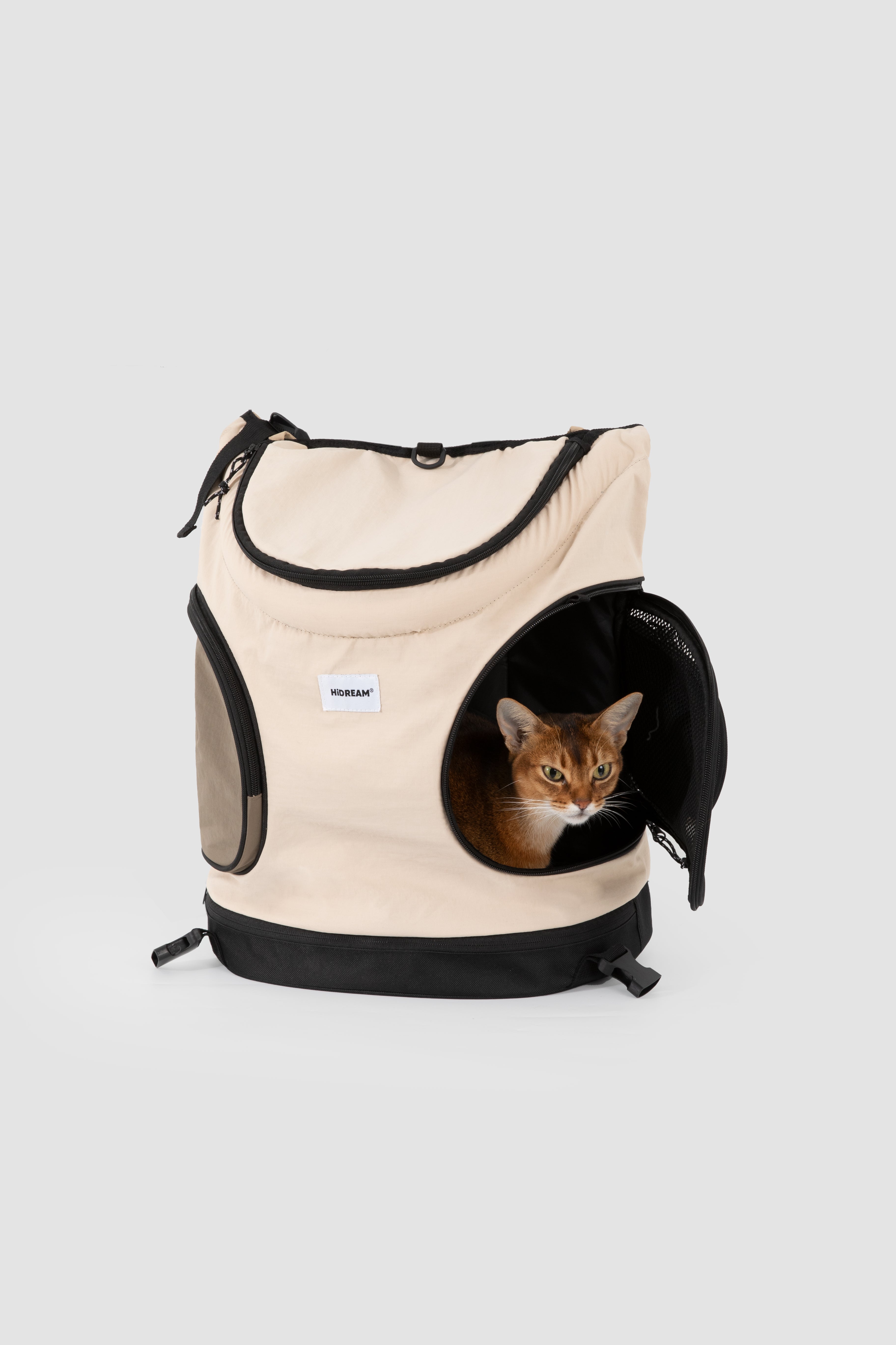 A beige and black pet backpack with a cat peeking out of its circular front opening, photographed on a light gray studio background.