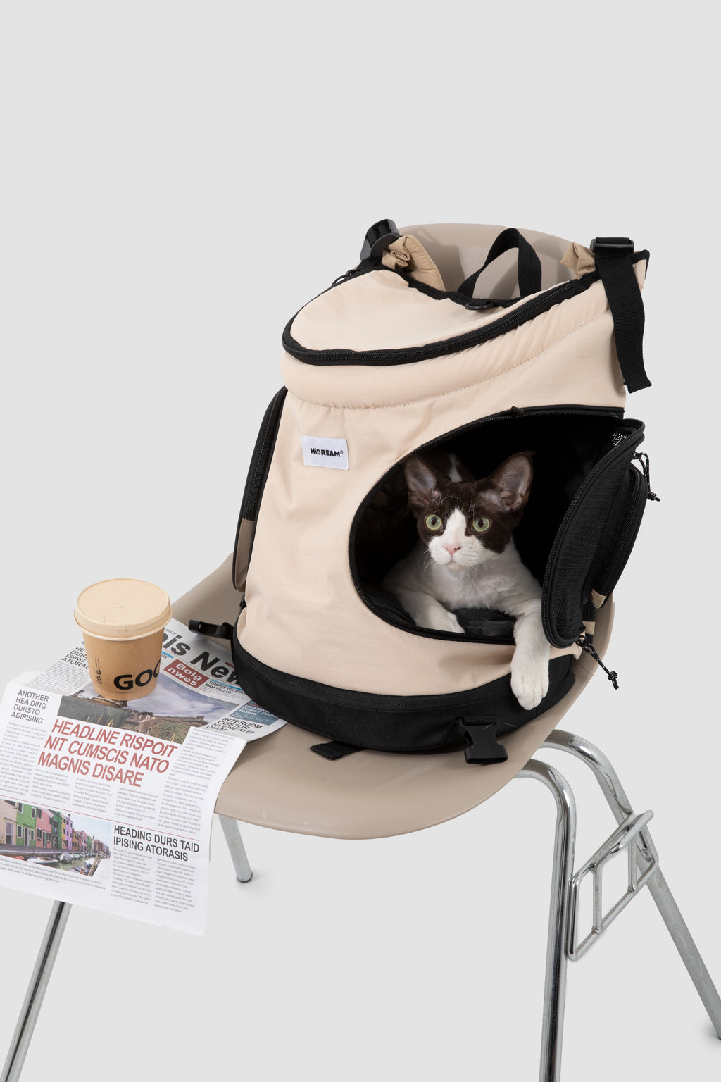 A beige and black pet carrier backpack with a cat peeking out of the front opening, styled on a chair next to a newspaper and coffee cup against a light gray background.