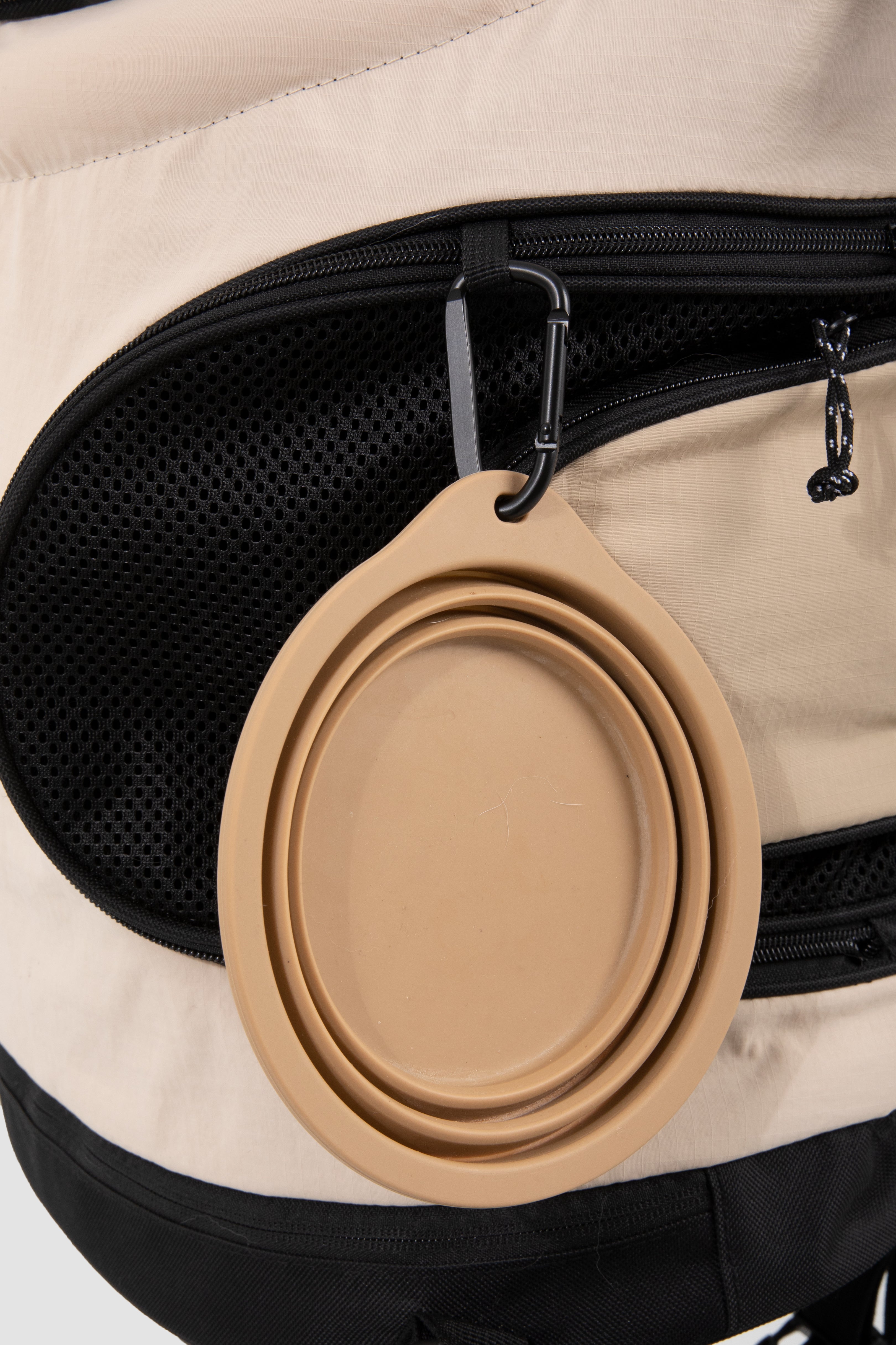 A beige collapsible silicone water bowl with a black carabiner clip, attached to a beige and black backpack.