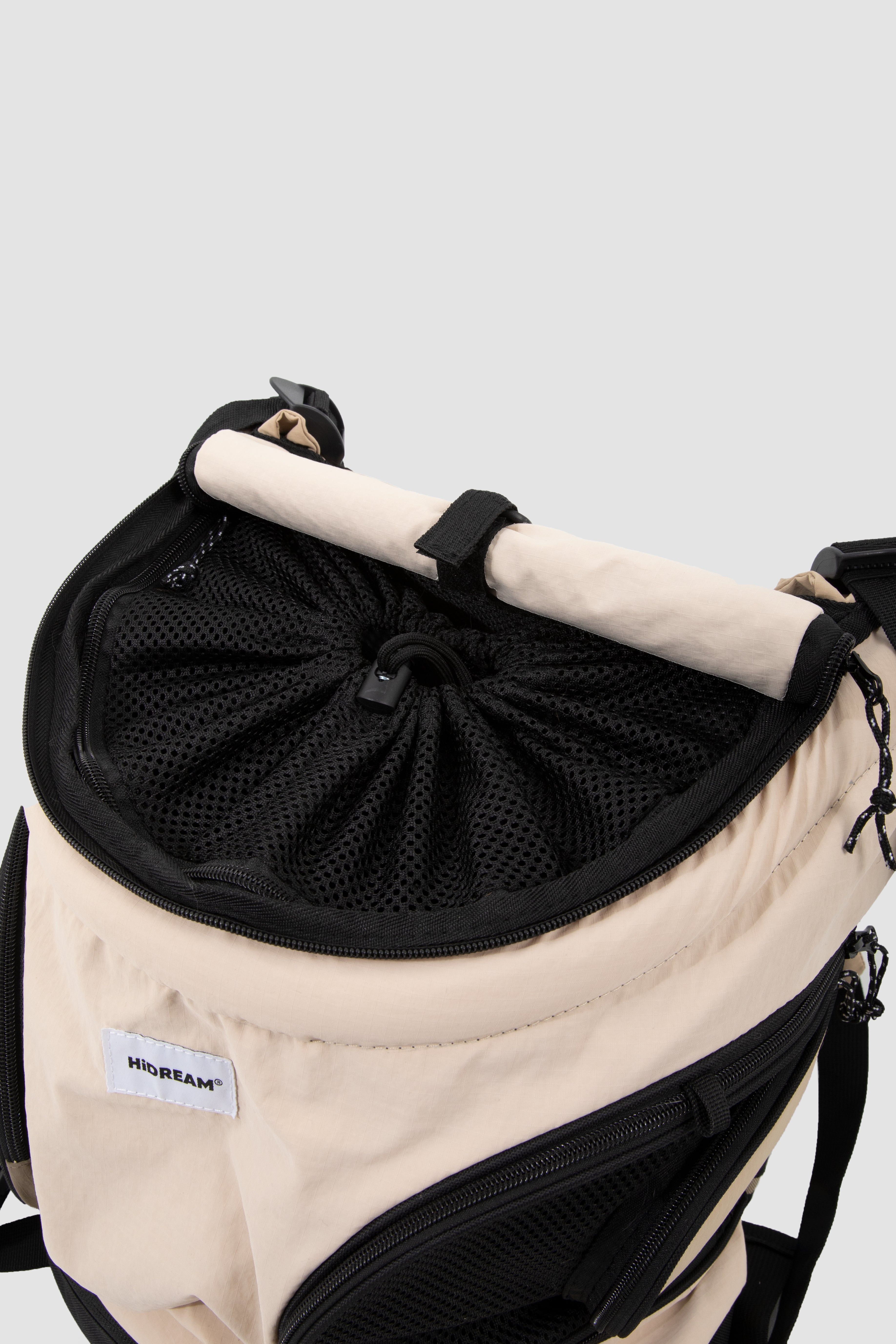 Close-up of a beige and black backpack showing its interior storage setup, including a black mesh drawstring compartment and a light beige rolled strap or handle, with a visible HIDEAM brand label.