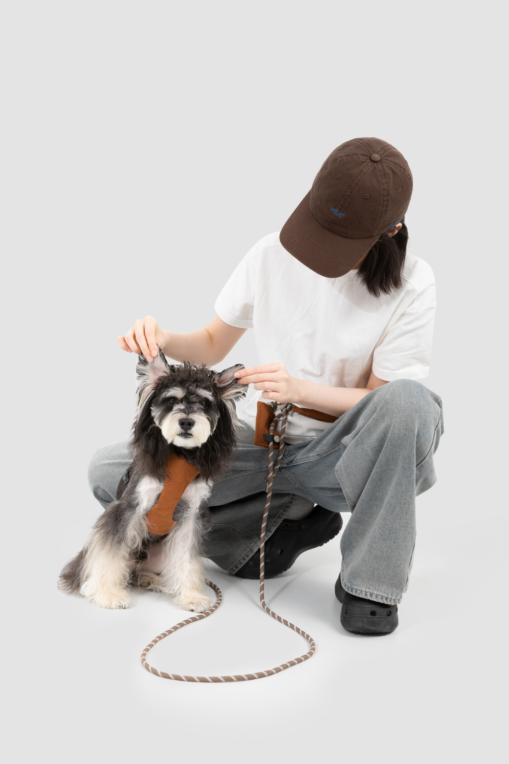 A person crouches beside a small gray and white dog wearing a brown harness, with the person holding the dog's head and the leash on a plain light gray studio background.