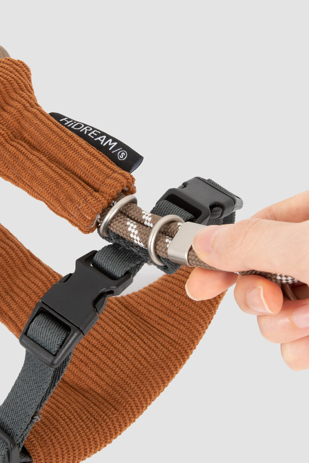 A close-up of a person's hand adjusting the metal buckle and textured strap of a brown ribbed dog harness, with a black HIDREAM brand tag visible.