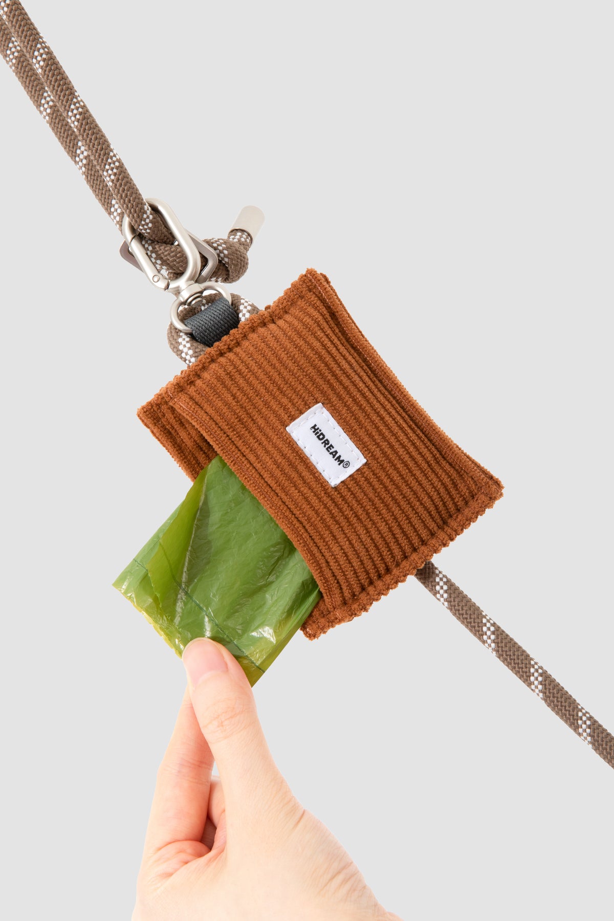 A close-up of a brown ribbed fabric dog leash pouch clipped to a brown leash, with a hand holding a green plastic bag behind the pouch.