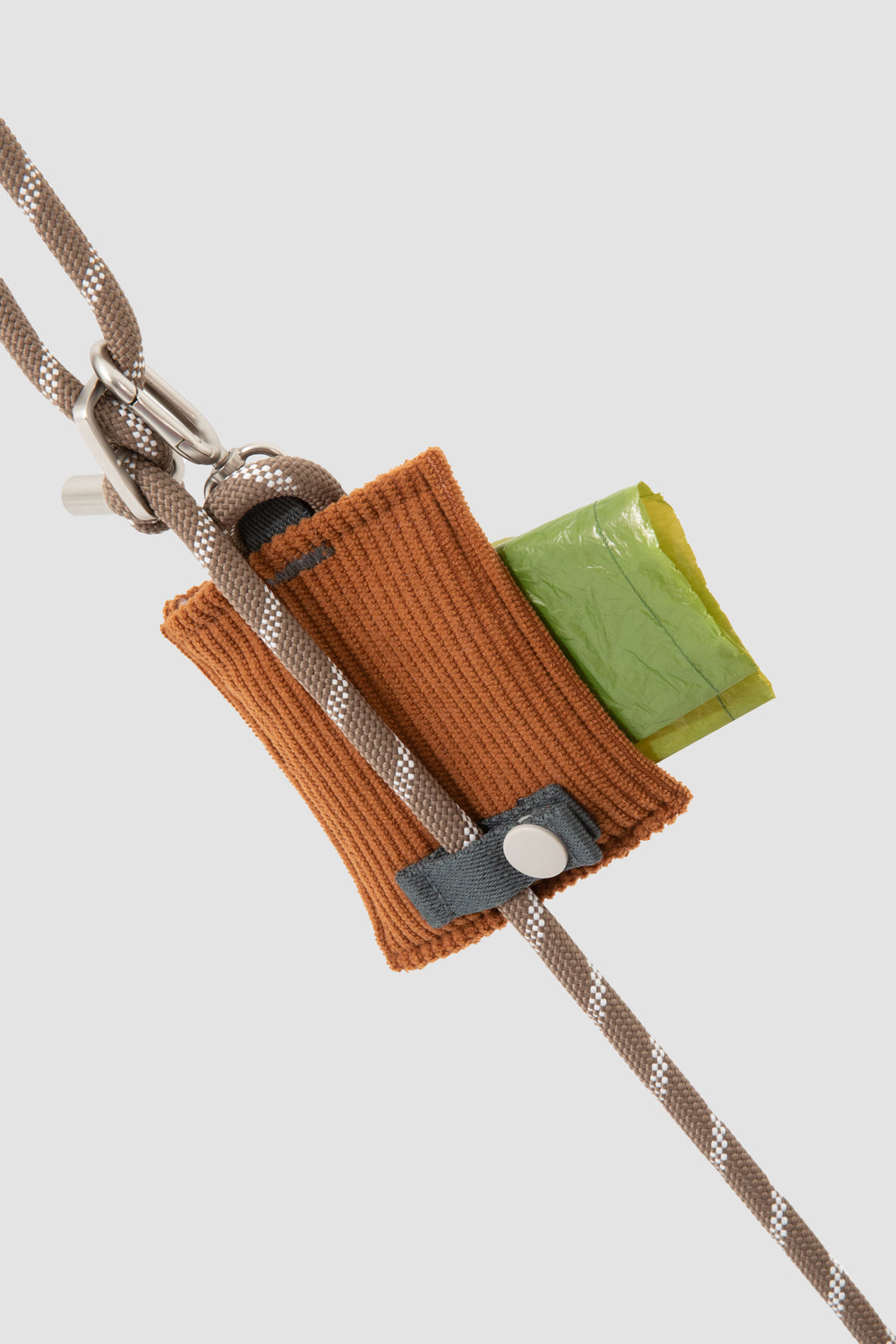 A close-up of a brown knitted pouch or loop attached to a braided rope, with a small green square piece and a gray tab, shown on a light gray background.