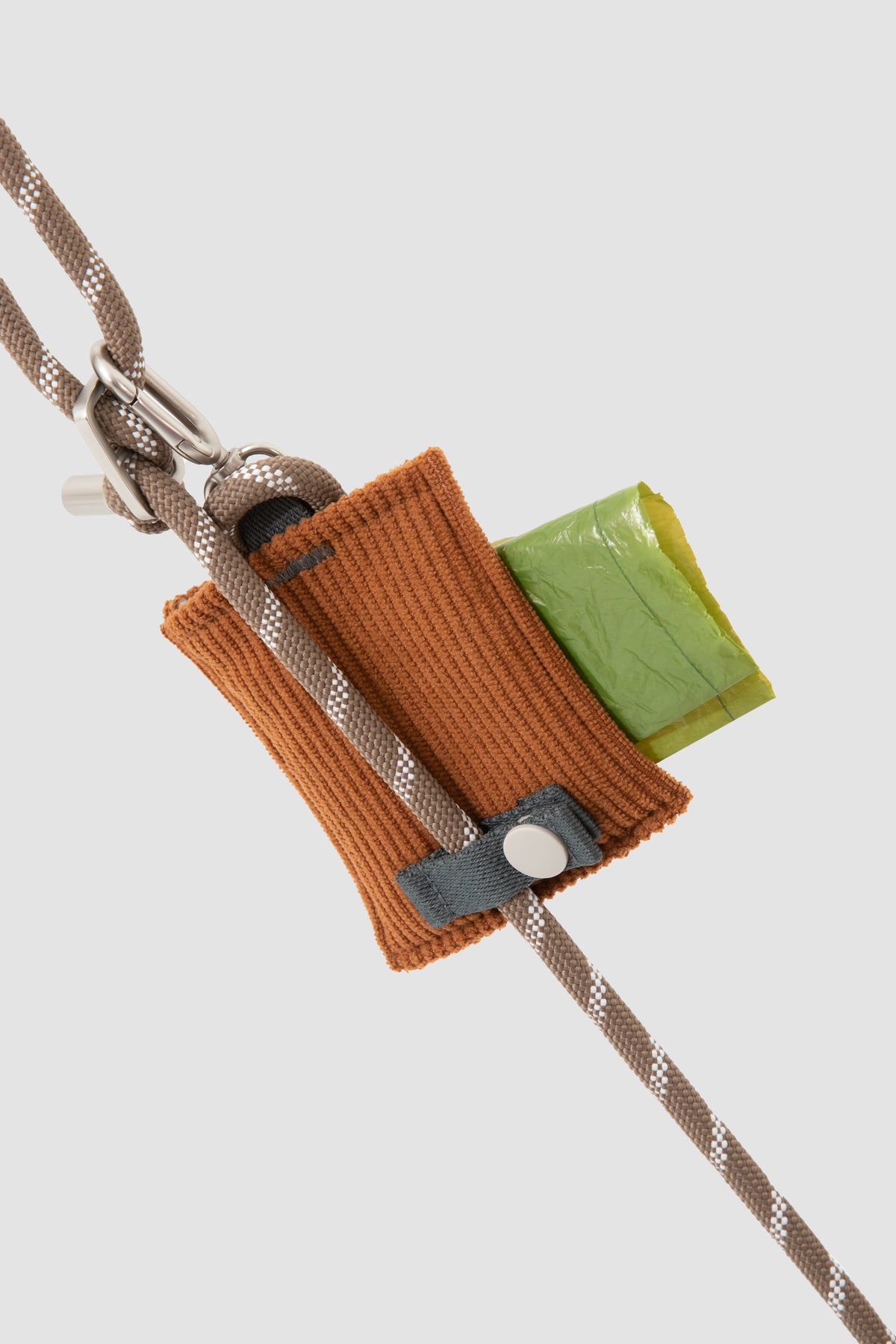 A close-up of a brown knitted pouch or loop attached to a braided rope, with a small green square piece and a gray tab, shown on a light gray background.
