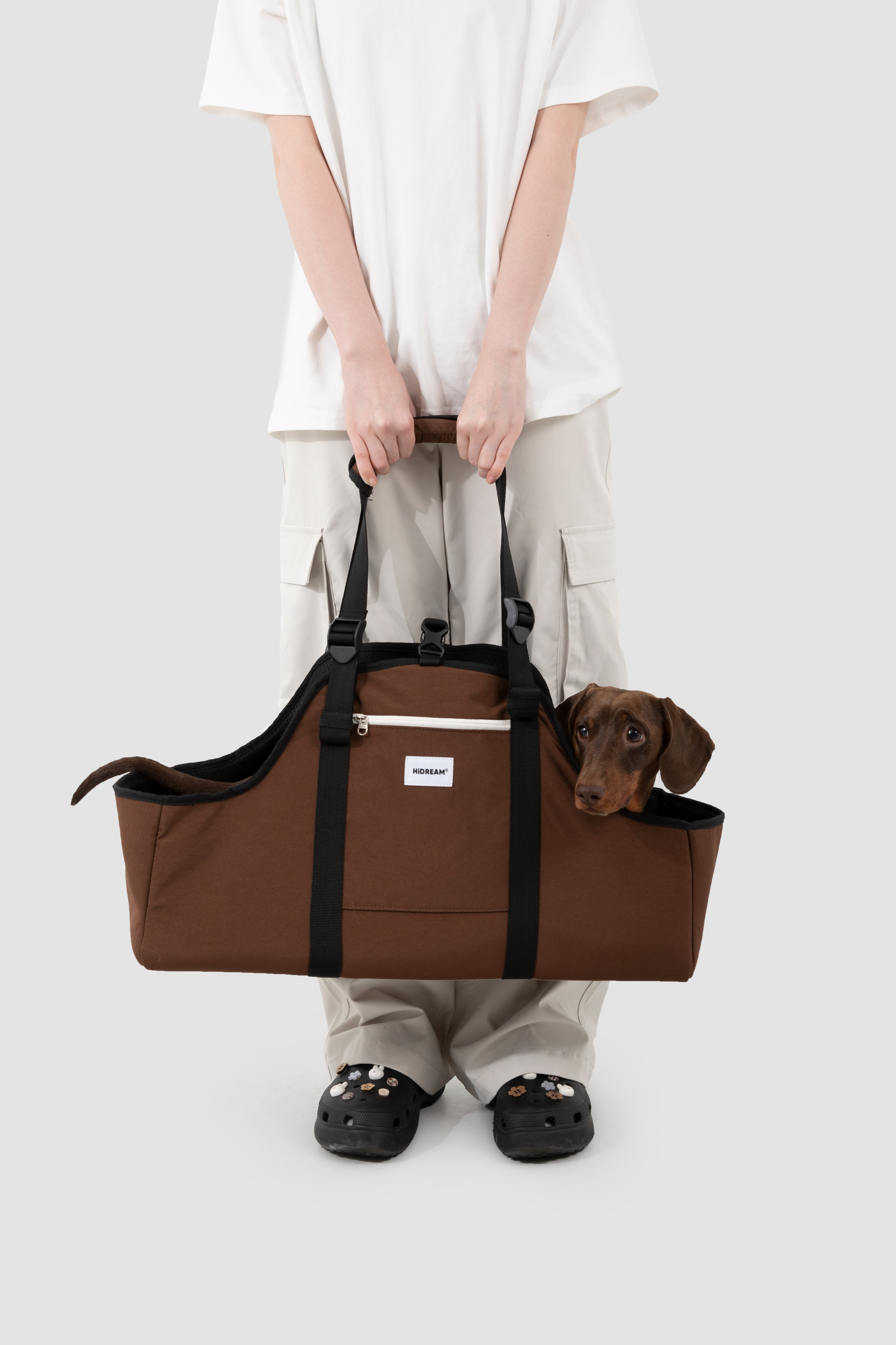 A person in a white t-shirt and beige cargo pants holds a brown pet carrier duffel bag with a small brown dog peeking out from the top, against a light gray studio background.