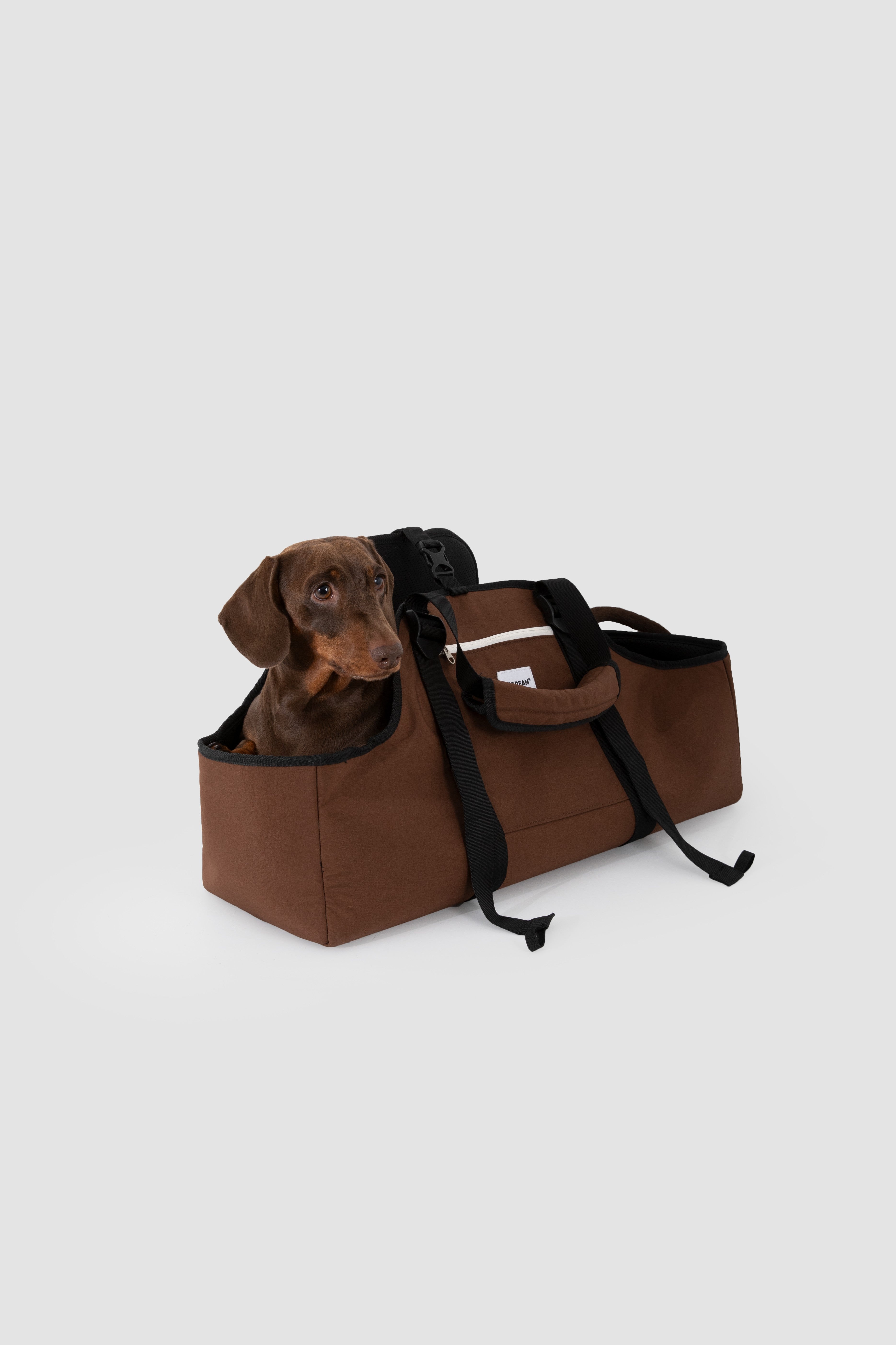 A brown dog carrier duffel bag with black straps, shown on a light gray background with a dachshund dog sitting inside the top opening.