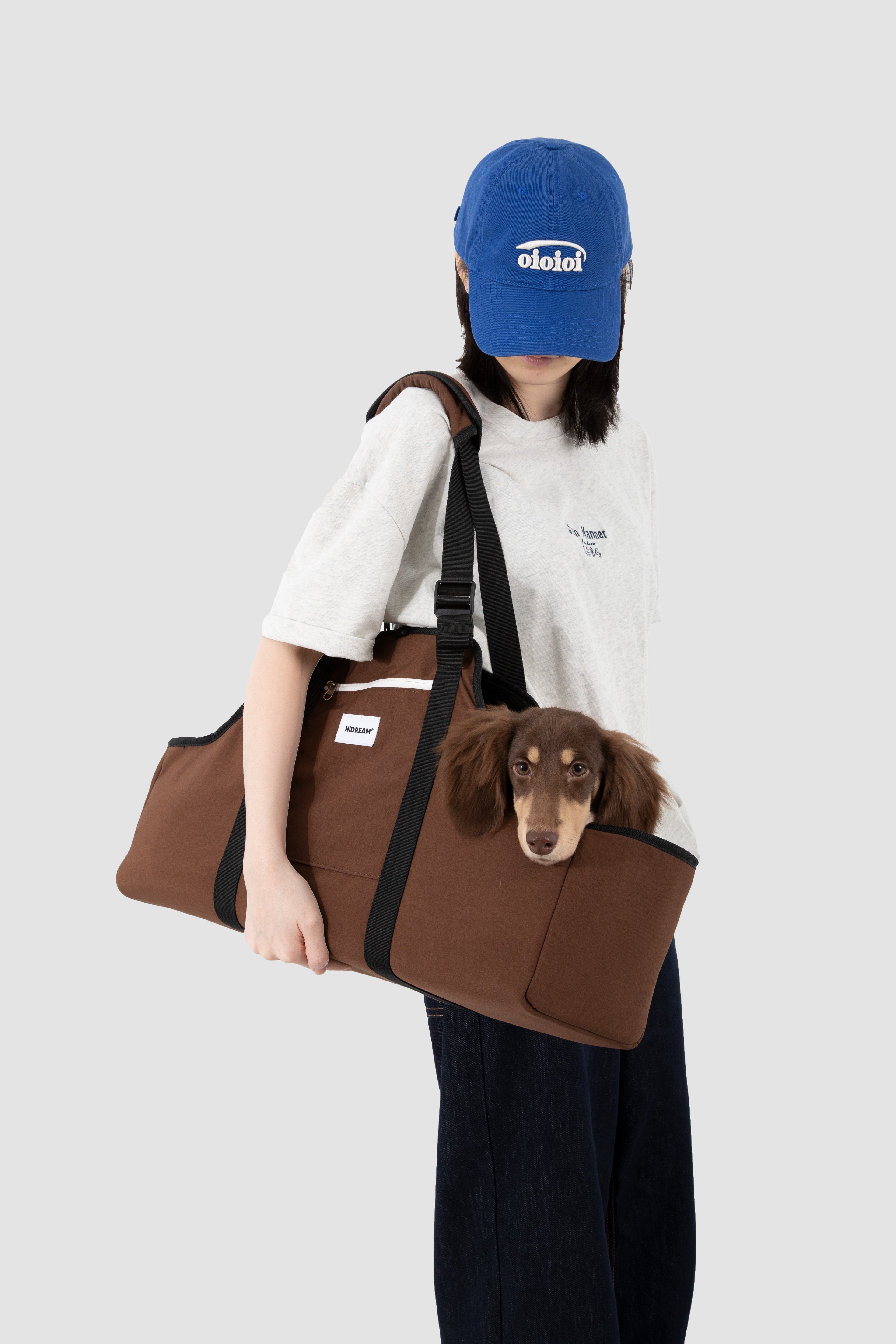 A person wearing a blue cap and white t-shirt carries a brown dachshund dog in a brown pet carrier tote bag with black straps, set against a solid light gray background.