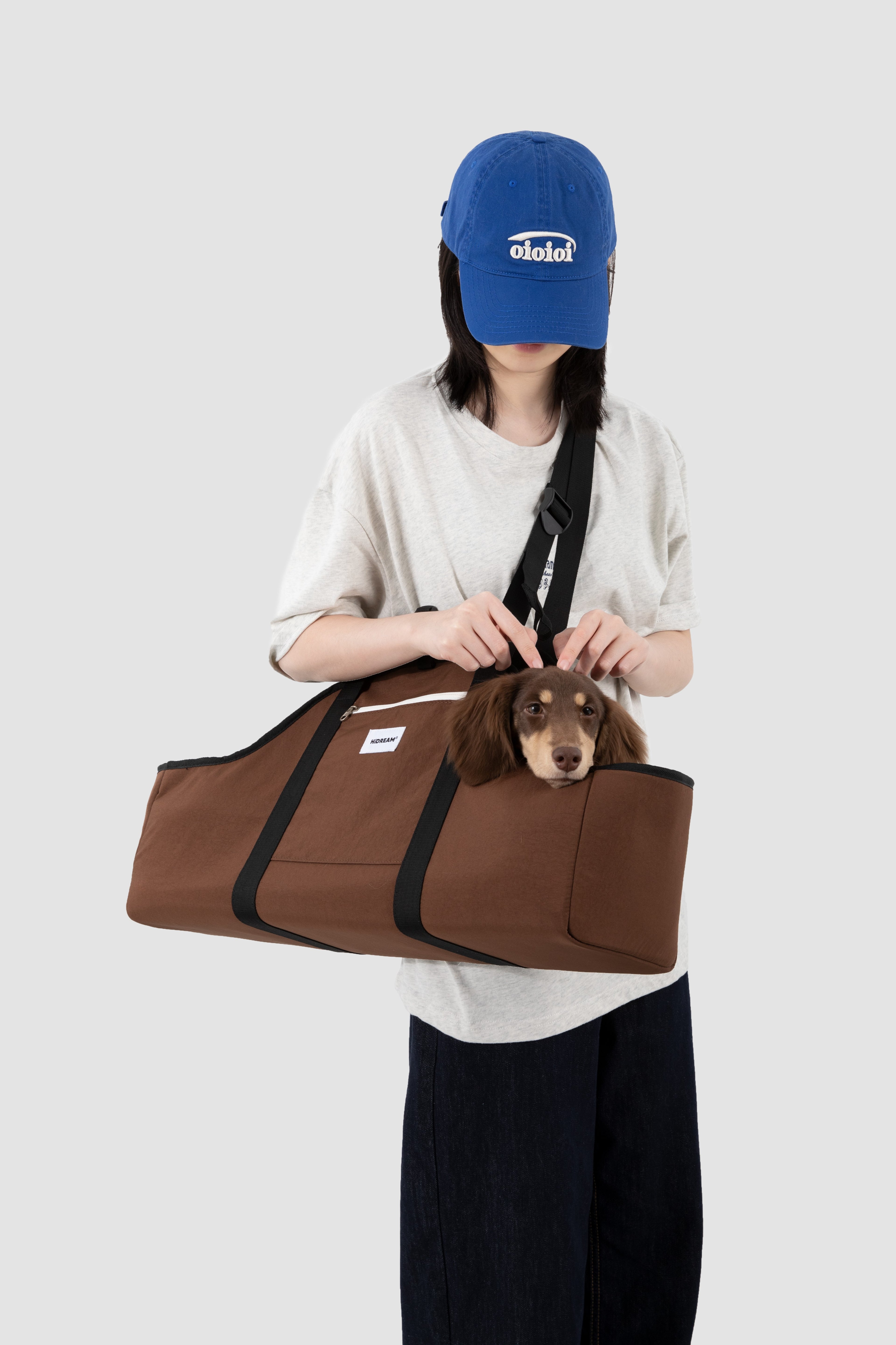 A person wearing a blue baseball cap and a light gray t-shirt holds a brown pet carrier bag with a small brown dachshund puppy peeking out. The bag has black straps and a white logo patch on the front, set against a solid light gray background.