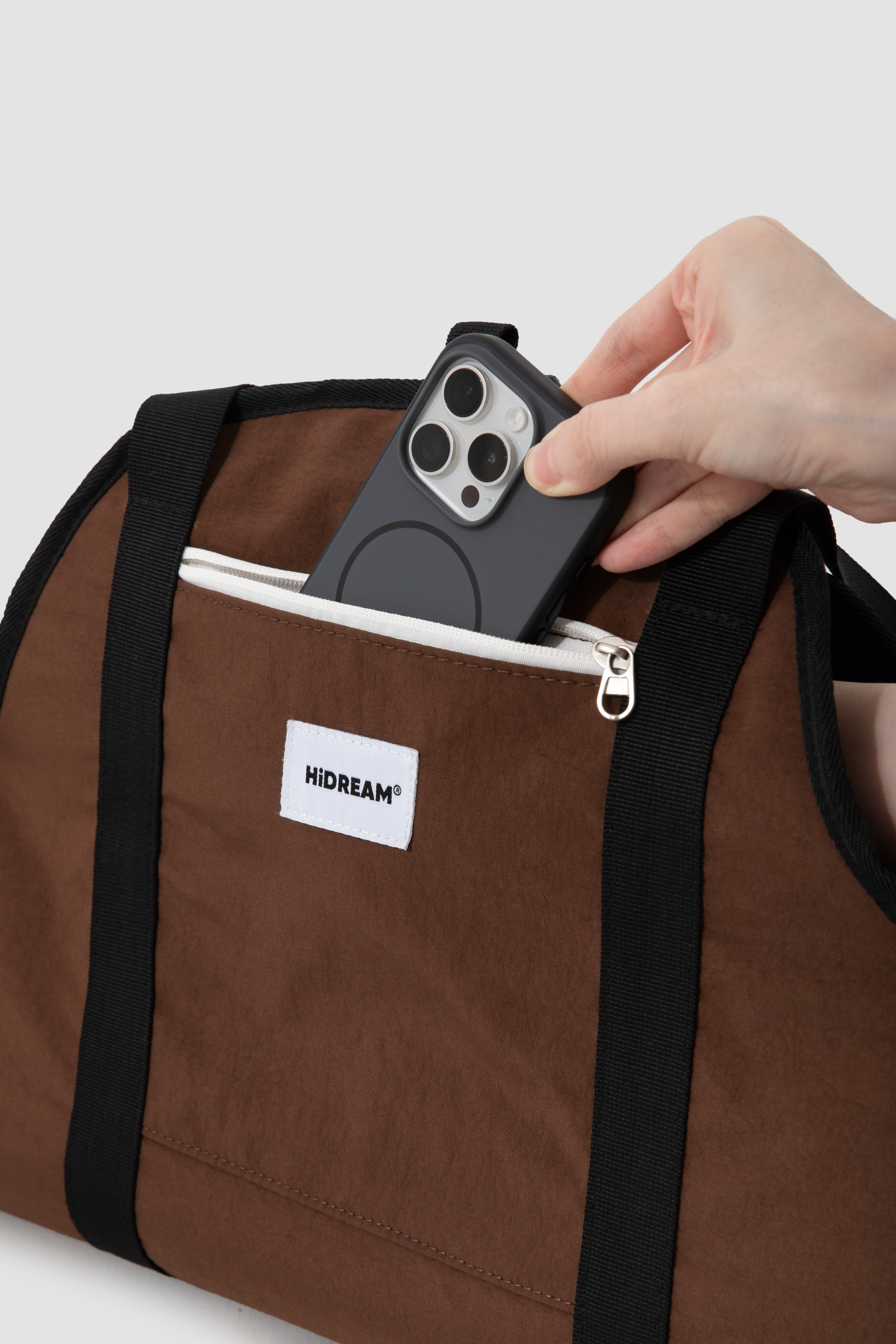 A close-up of a brown and black Duffle bag with a hand inserting a black smartphone into its front zippered pocket. The bag features a white brand label that reads 'HIDREAM'.