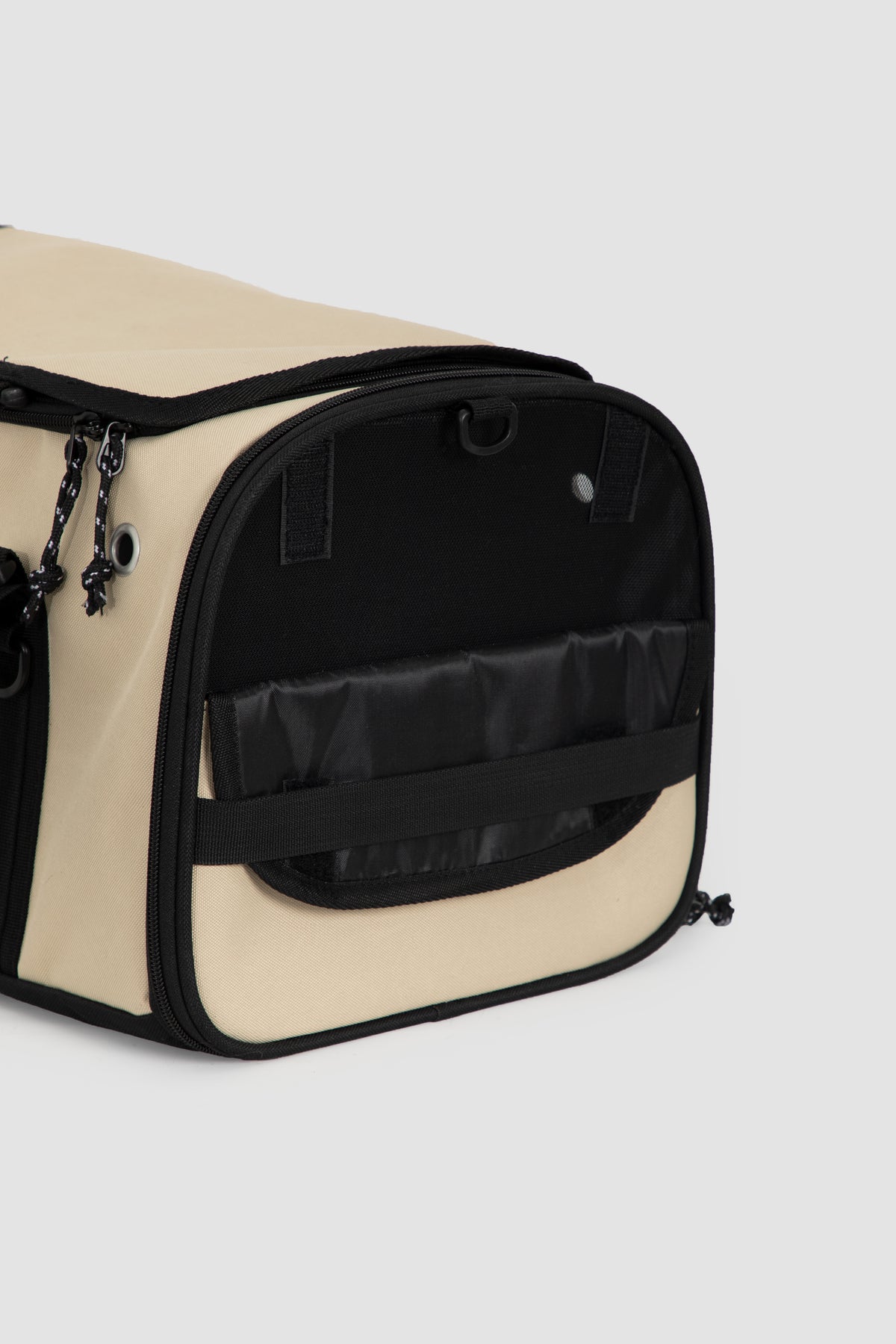 A close-up of a beige and black pet carrier or duffel bag with a front mesh window and black trim, shown on a light gray background.