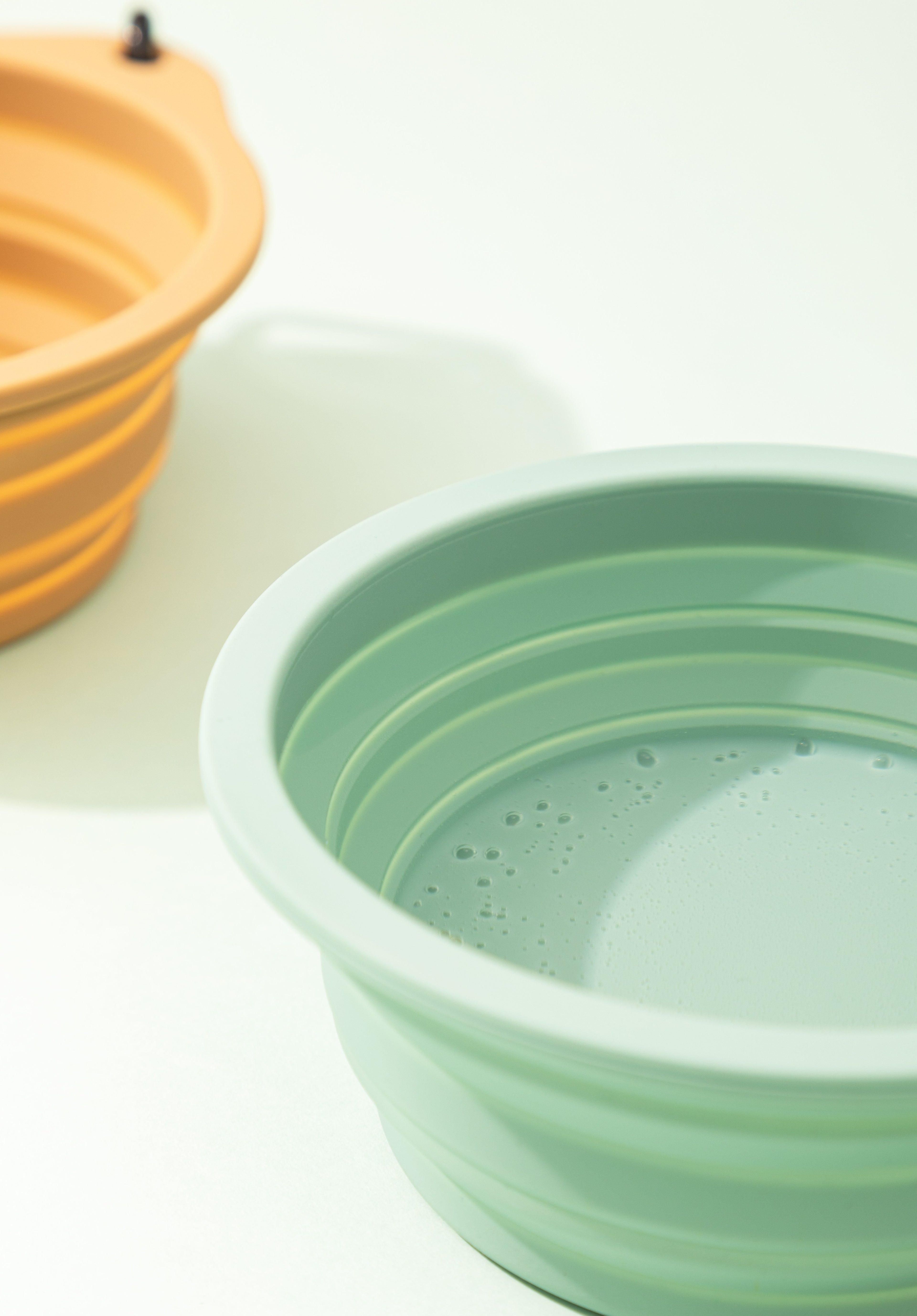 A mint green collapsible silicone bowl in the foreground on a white surface, with a second orange-brown bowl blurred in the background.