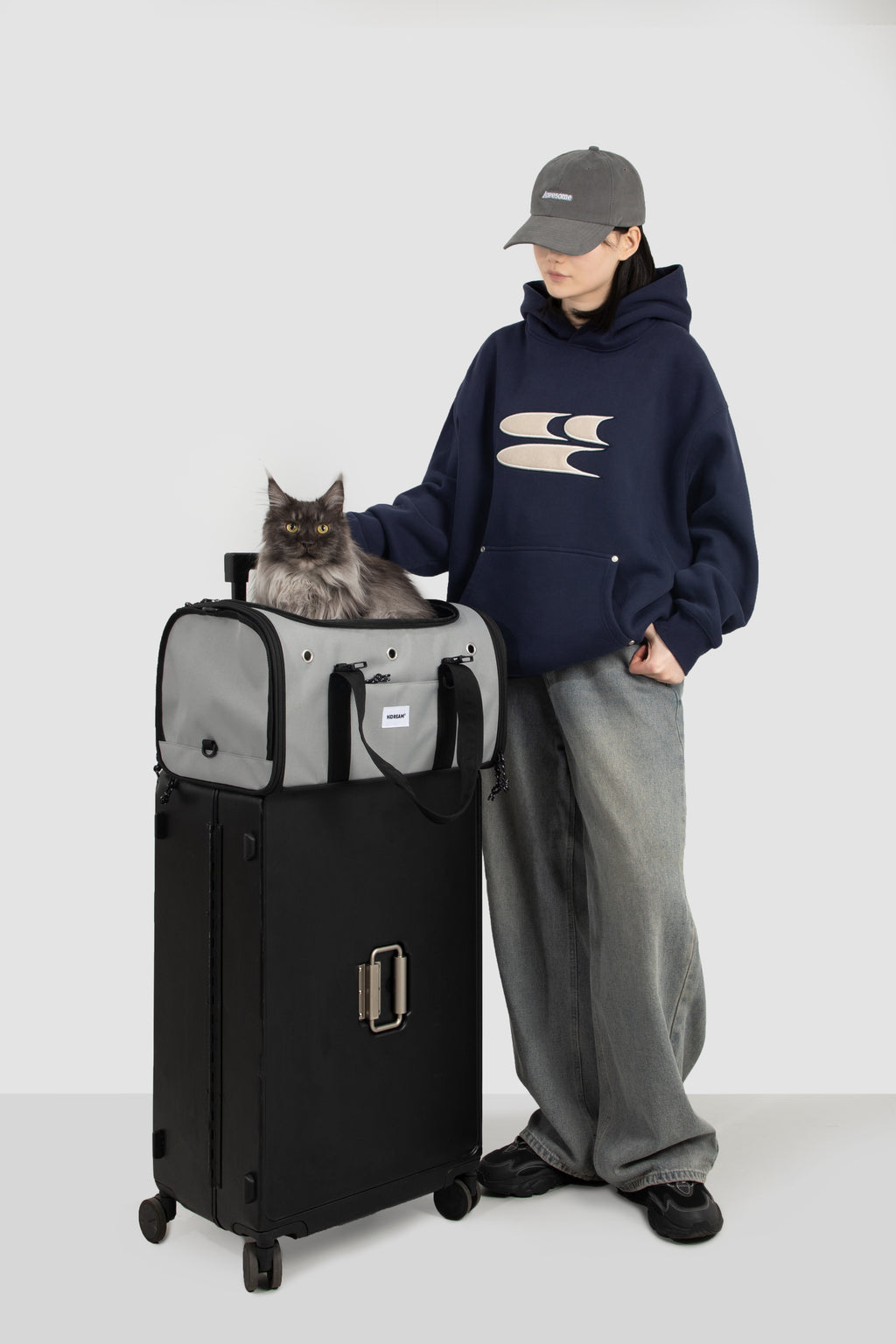 A person wearing a navy hoodie, gray sweatpants, and a gray cap stands next to a black and gray pet suitcase with a gray cat sitting on top, against a light gray studio background.