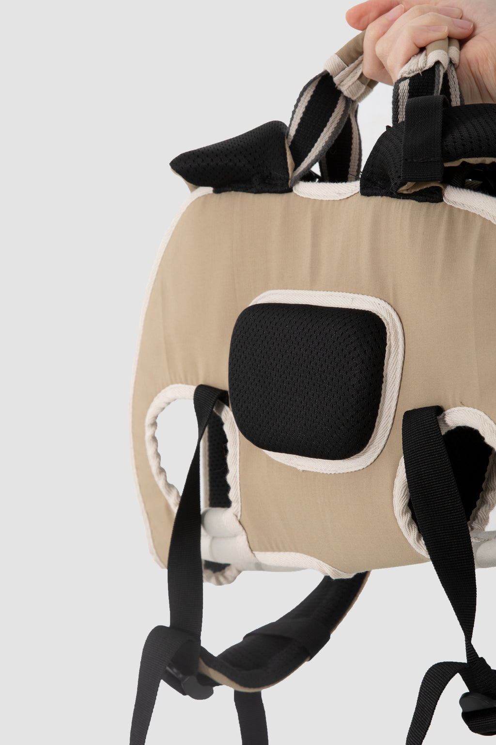 A close-up of a beige baby carrier or backpack-style baby support worn on a person's shoulder, with black padded mesh panels, white trim, and a hand holding the top handle against a light gray background.