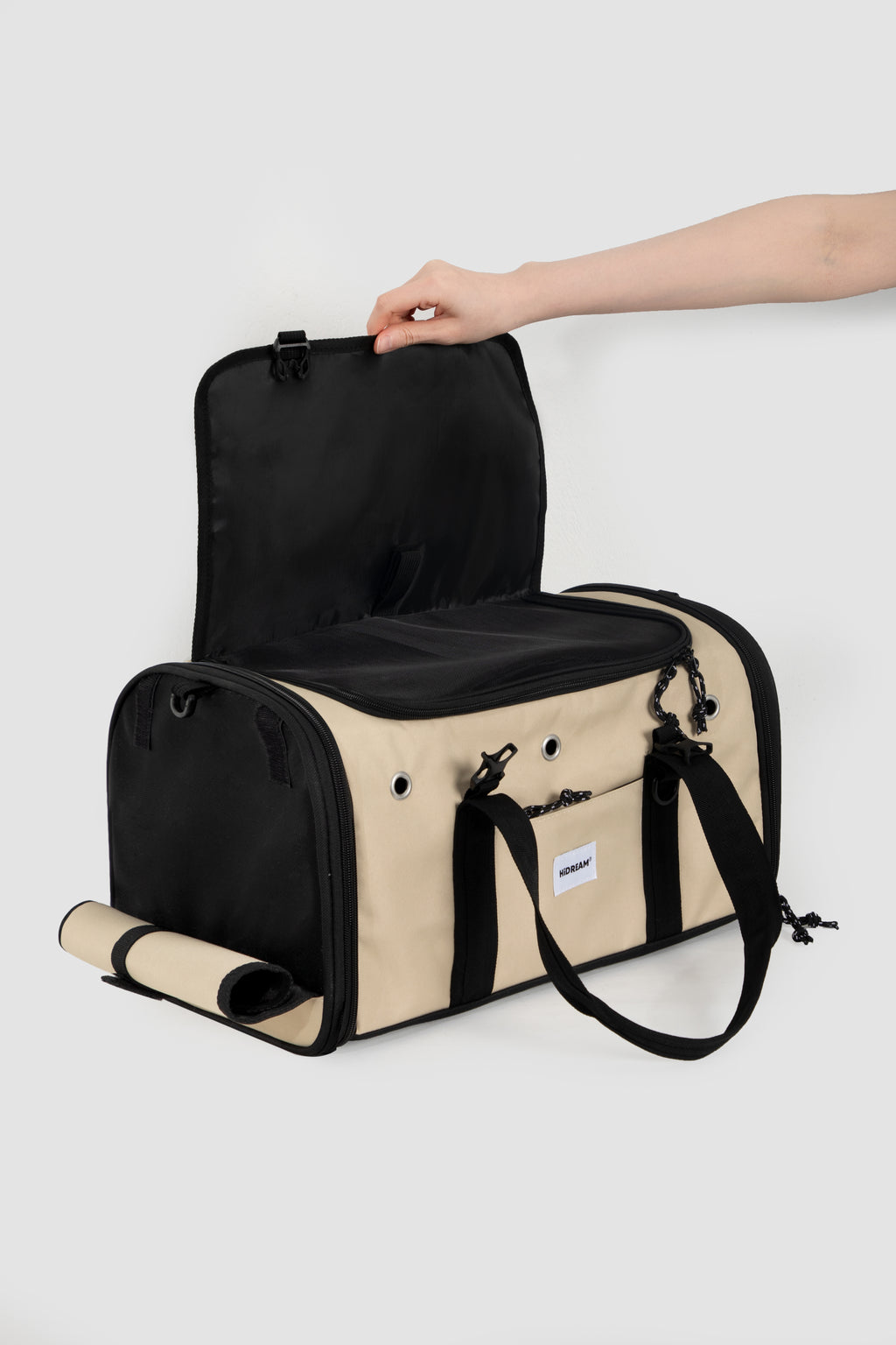 A beige and black duffel bag with a hand lifting the main compartment lid, shown on a light gray background.