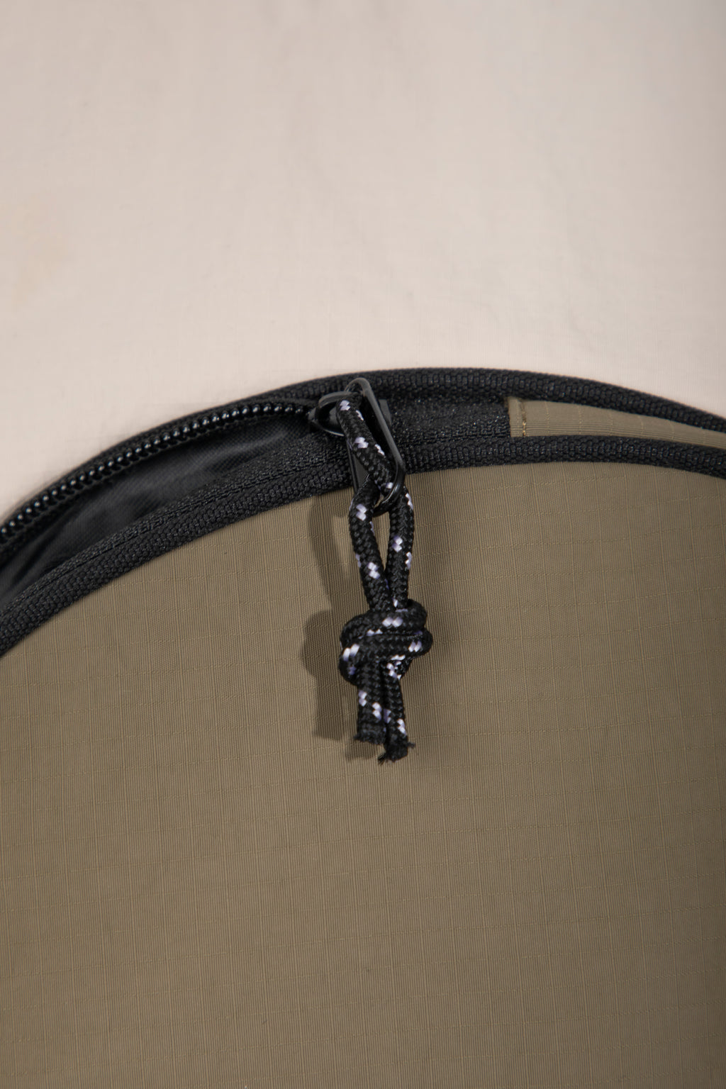 Close-up detail of a circular or dome-shaped product, showing a black zipper with a knotted black-and-white cord pull tab against a warm beige background.