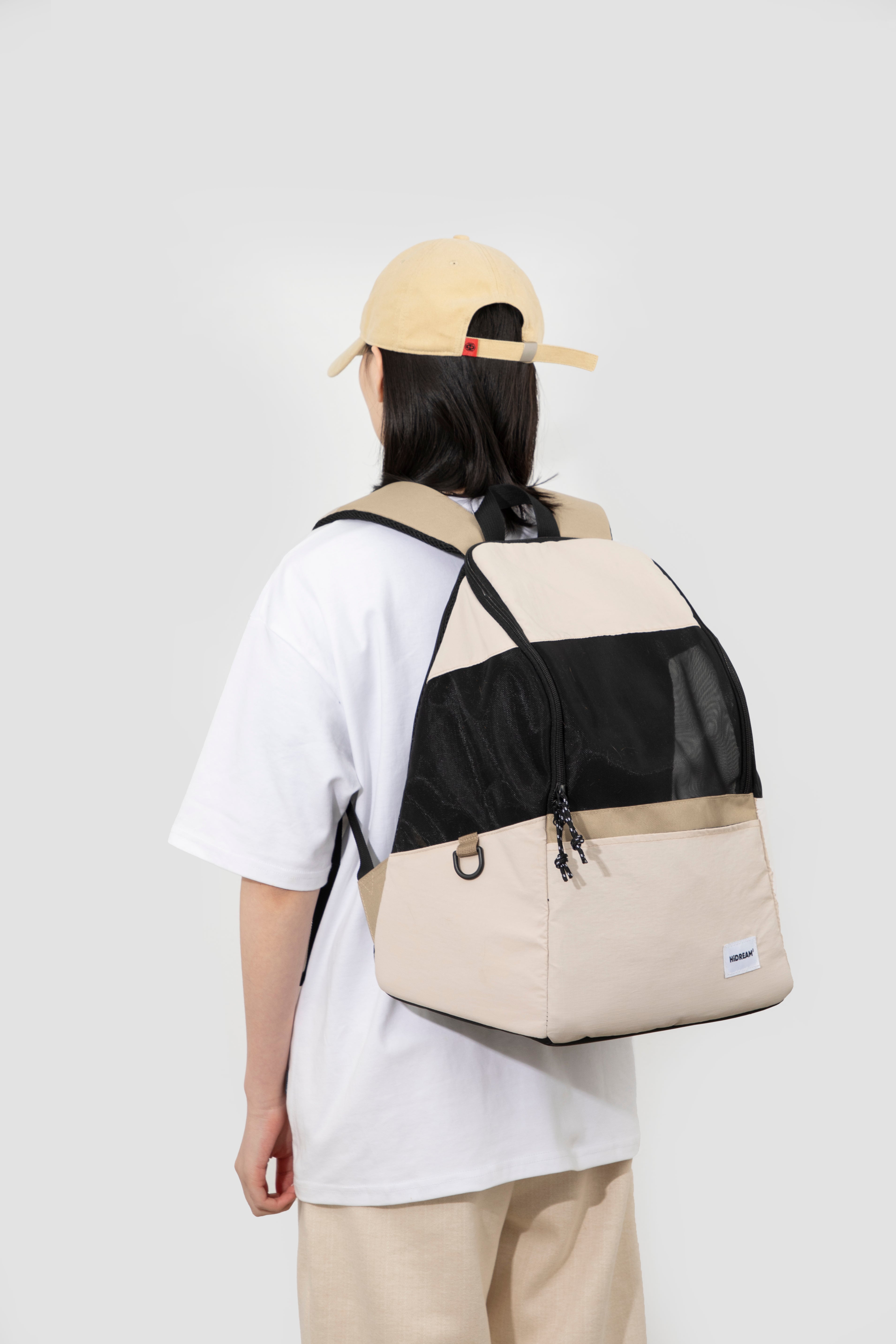 A person shown from the back wearing a beige and black color-blocked backpack, a white t-shirt, and a beige baseball cap against a light gray studio background.