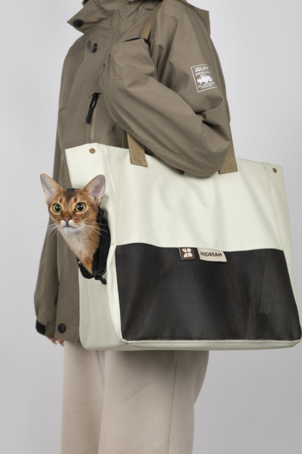 A person wearing a brown jacket and beige pants carries a large off-white and black pet tote bag with a cat peeking out from the side pocket, against a light gray studio background.