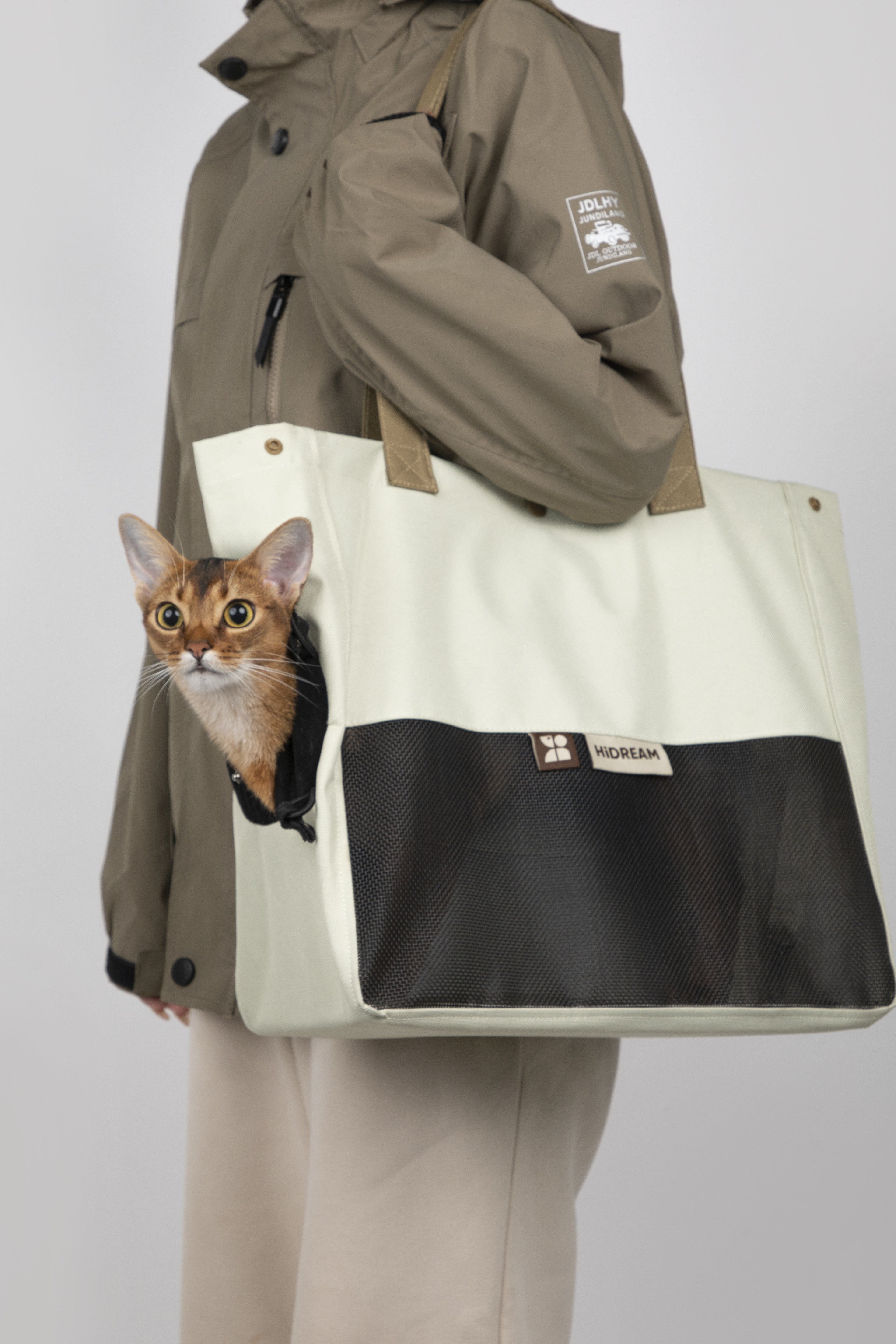A person wearing a brown jacket and beige pants carries a large off-white and black pet tote bag with a cat peeking out from the side pocket, against a light gray studio background.