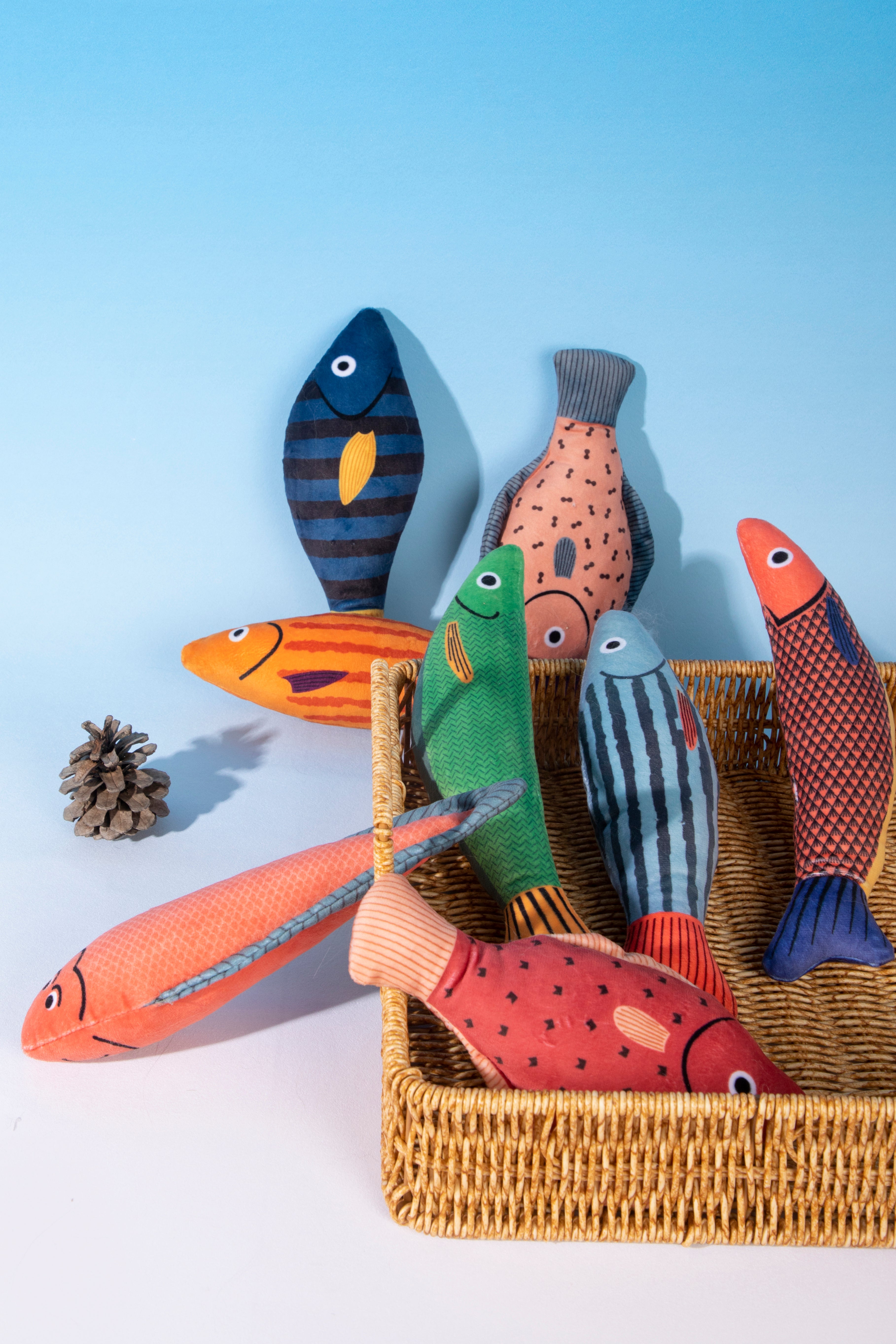 A set of colorful handmade fabric fish toys arranged on and around a woven wicker basket, with a small pinecone prop on a light blue and white background.