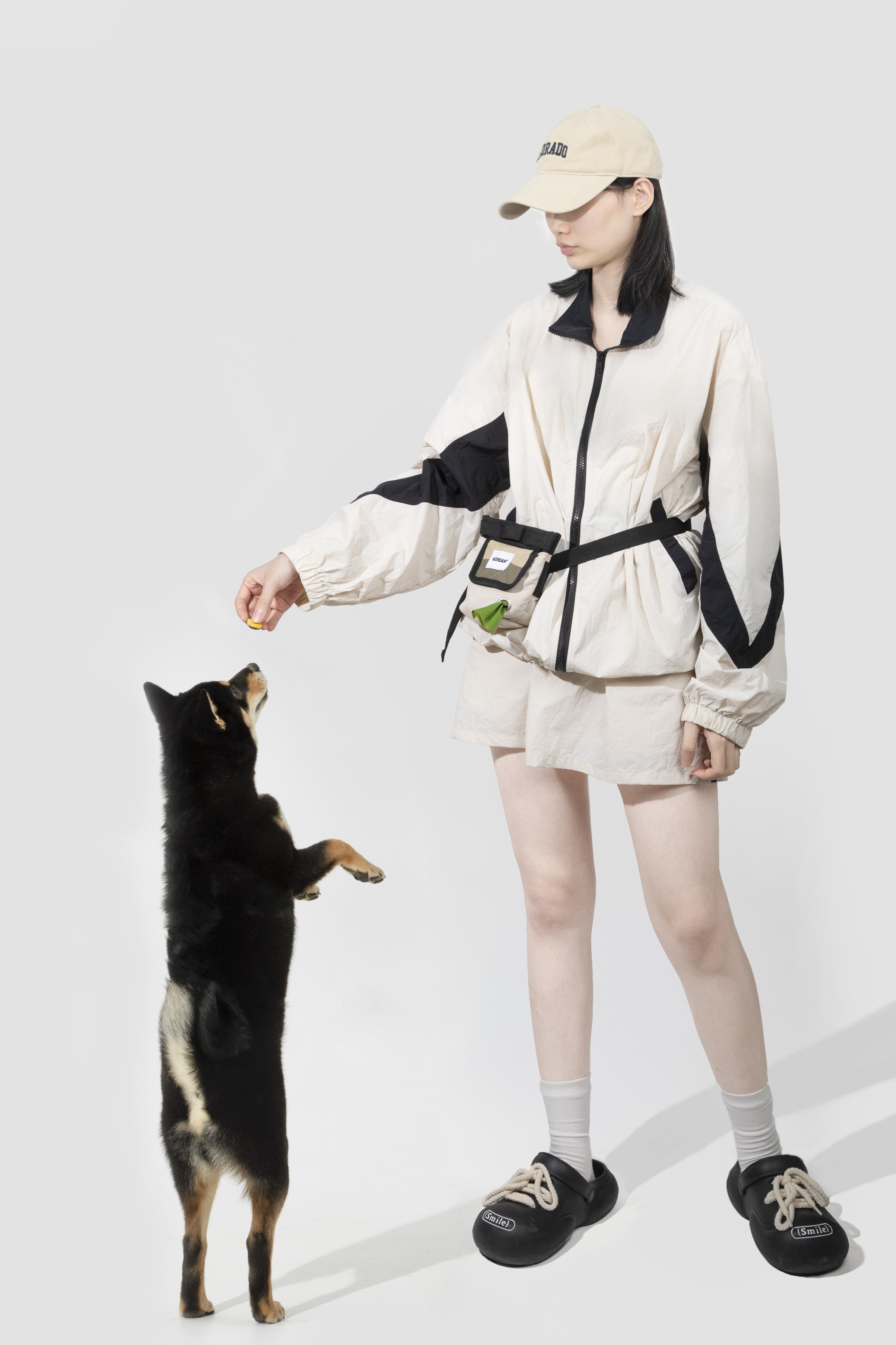 A woman wearing a beige and black windbreaker jacket, matching shorts, a beige cap, and black sneakers stands on a light gray studio background, reaching down to a small black and tan dog standing on its hind legs.
