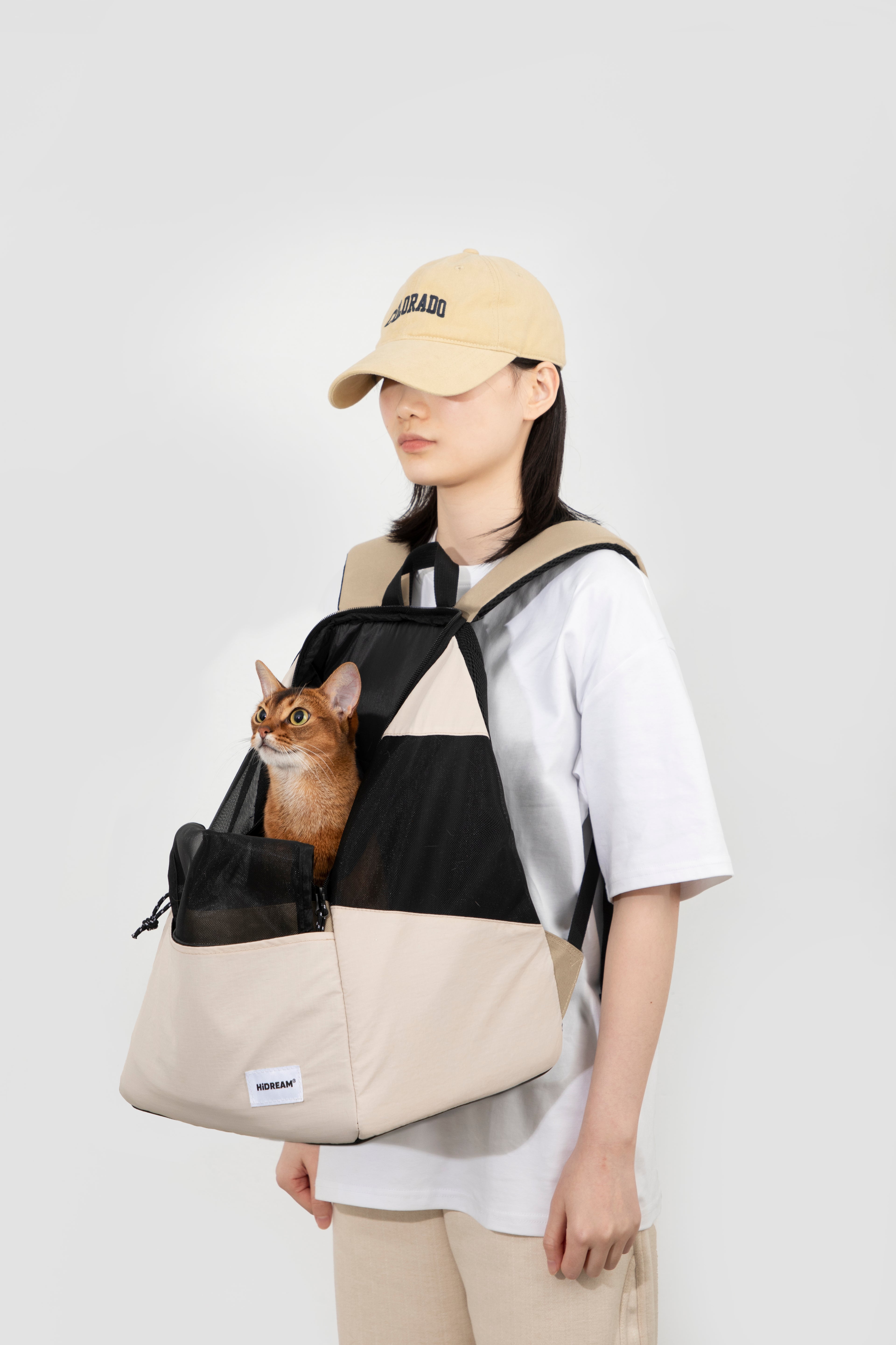 A person wearing a beige cap, white t-shirt, and beige pants carries a brown cat in a beige and black pet backpack carrier, set against a plain light gray studio background.