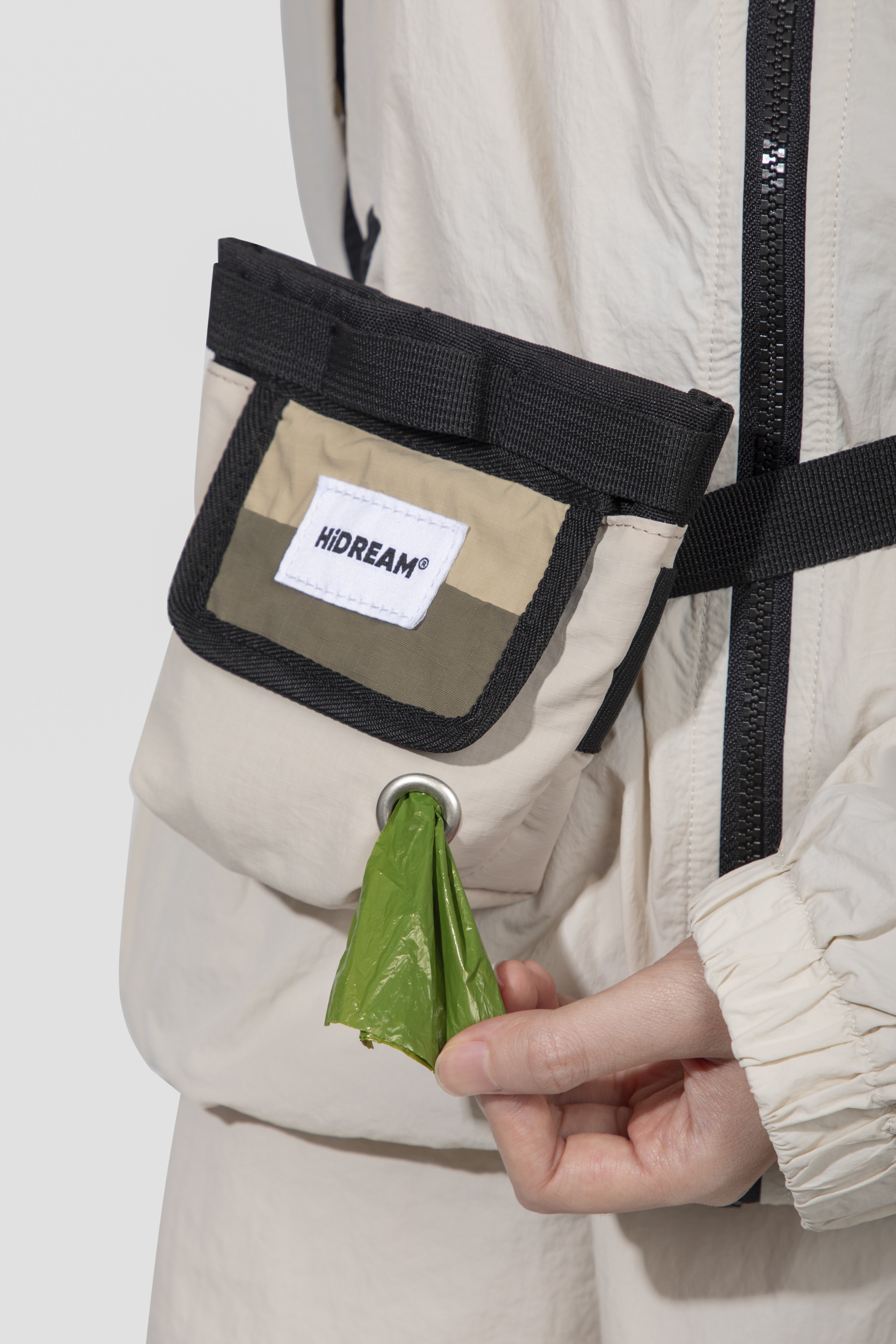 Close-up of a person wearing a beige and black utility pouch on their jacket, with a green plastic bag inserted into the pouch's ring, and a visible HIDREAM brand label.