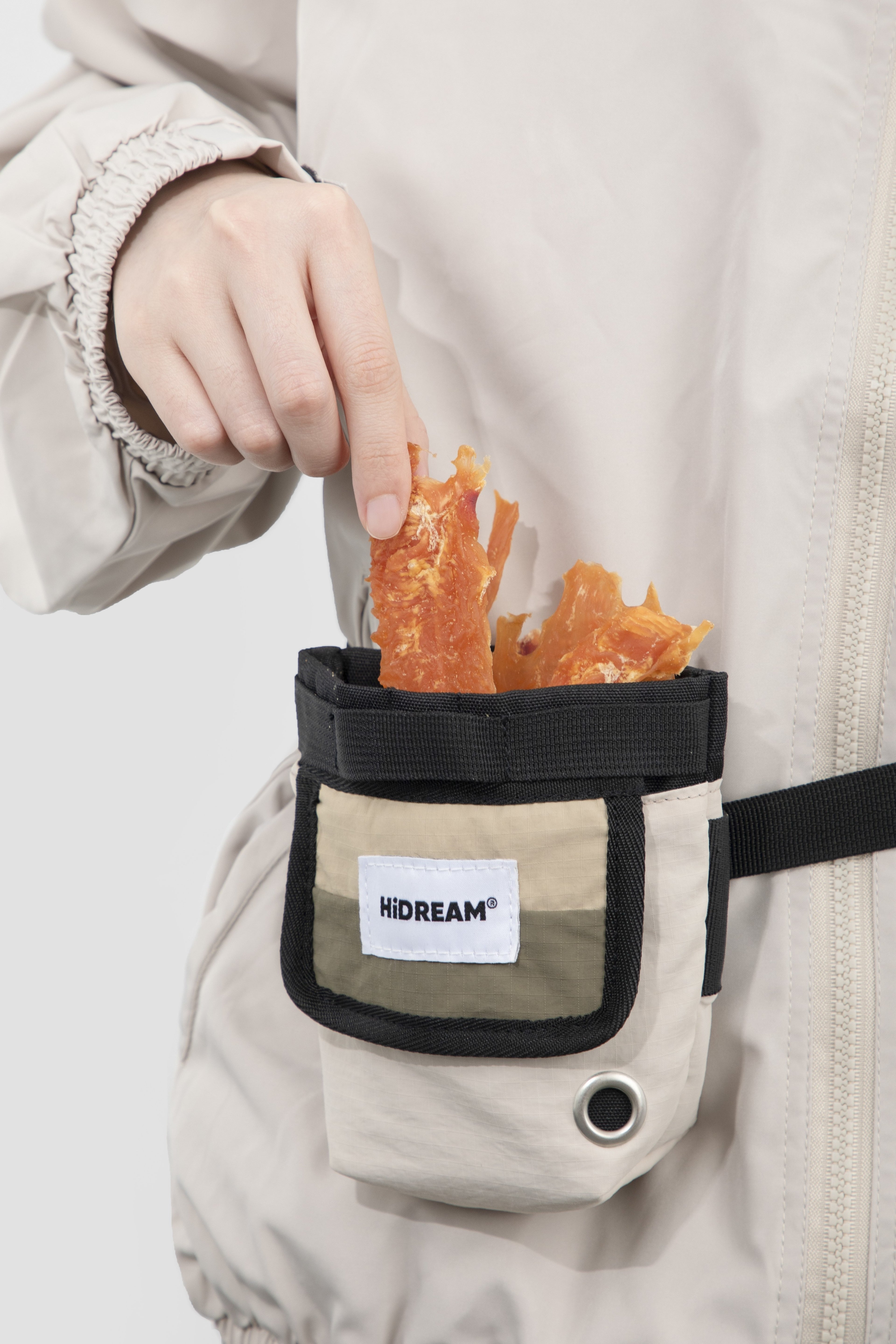 A close-up of a person wearing a light beige jacket and holding a small front pouch filled with orange dried fish or seaweed. The pouch has a black strap, black trim, and a white label reading HIDREAM.