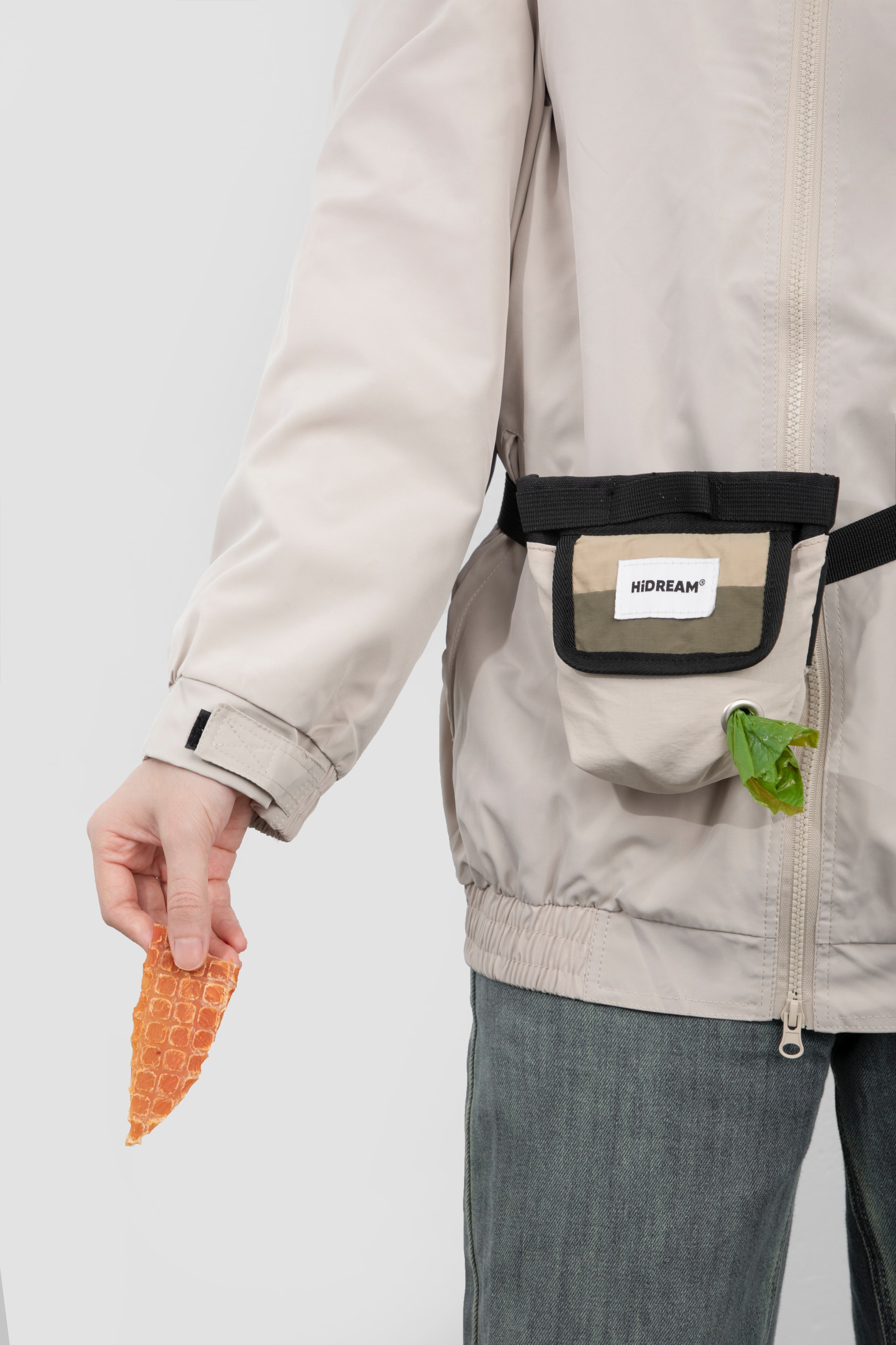 A close-up of a person wearing a light beige jacket with a black and beige front utility pouch. The pouch has a white label reading 'HIDREAM' and a small green rubbish bag is tucked into the side pocket.