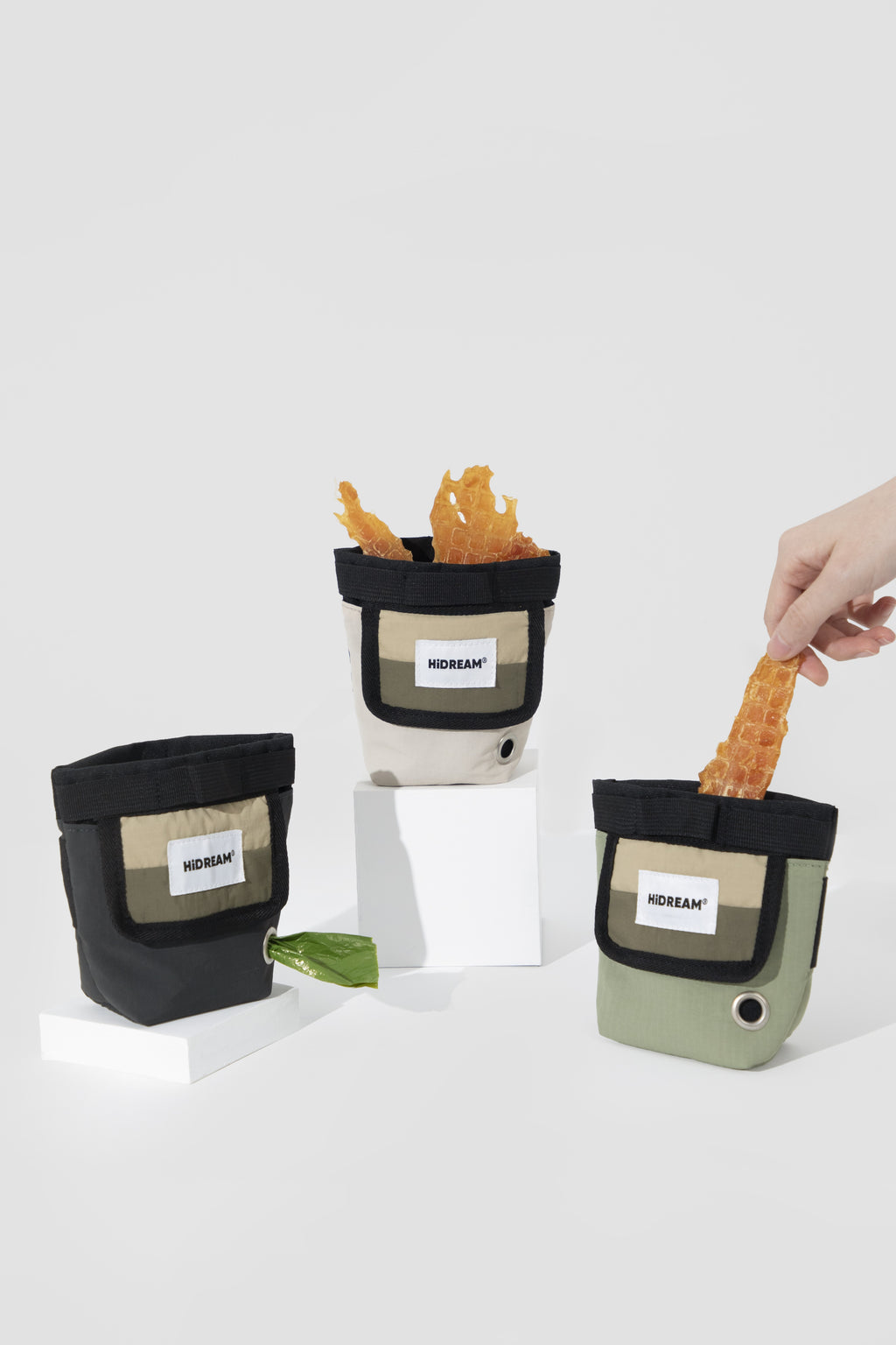 Three small pouch-style containers with visible HiDREAM labels are arranged on white blocks against a light gray studio background. Two containers are empty, one is filled with orange chips, and a human hand is reaching in from the right to take a chip.