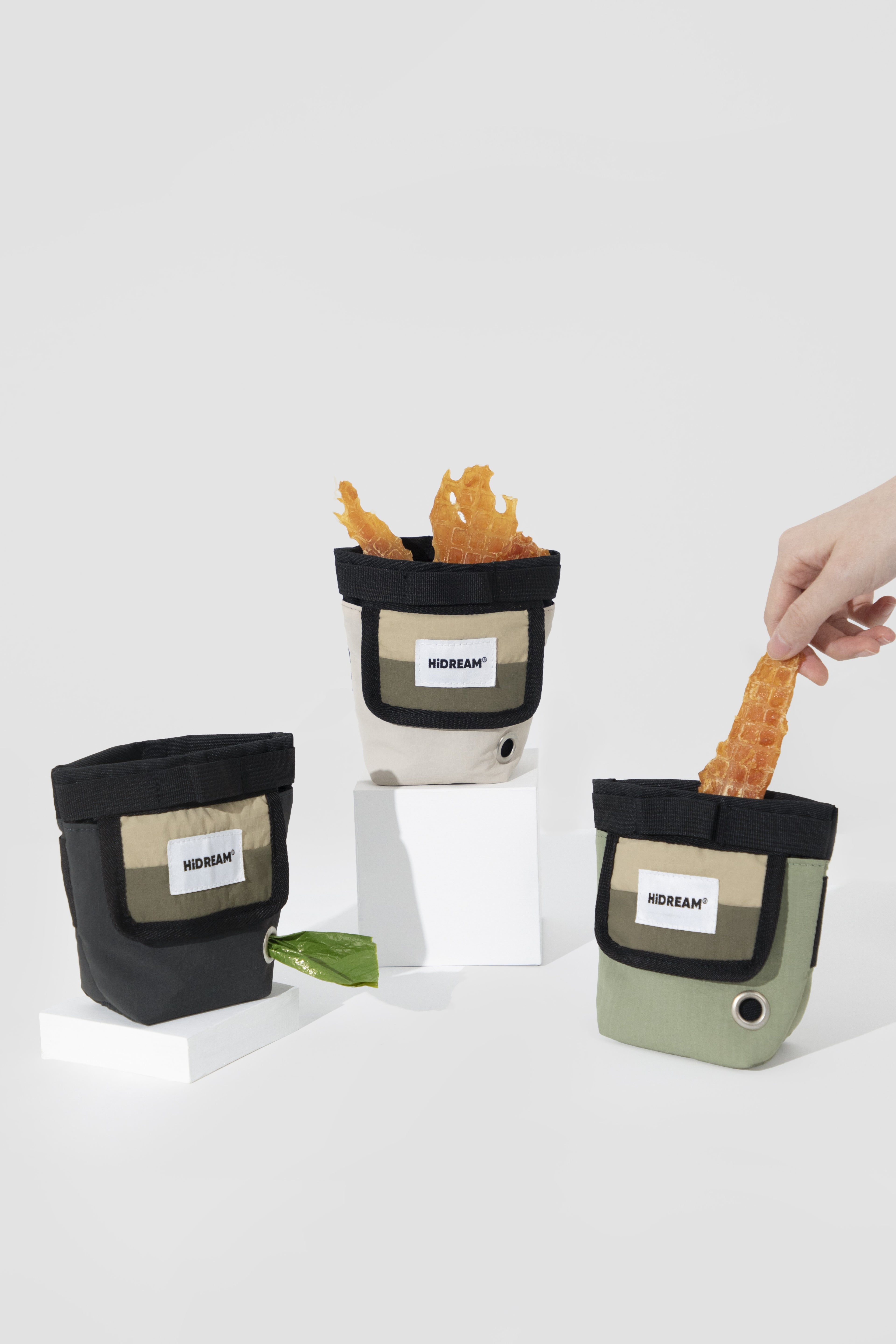 Three small pouch-style containers with visible HiDREAM labels are arranged on white blocks against a light gray studio background. Two containers are empty, one is filled with orange chips, and a human hand is reaching in from the right to take a chip.