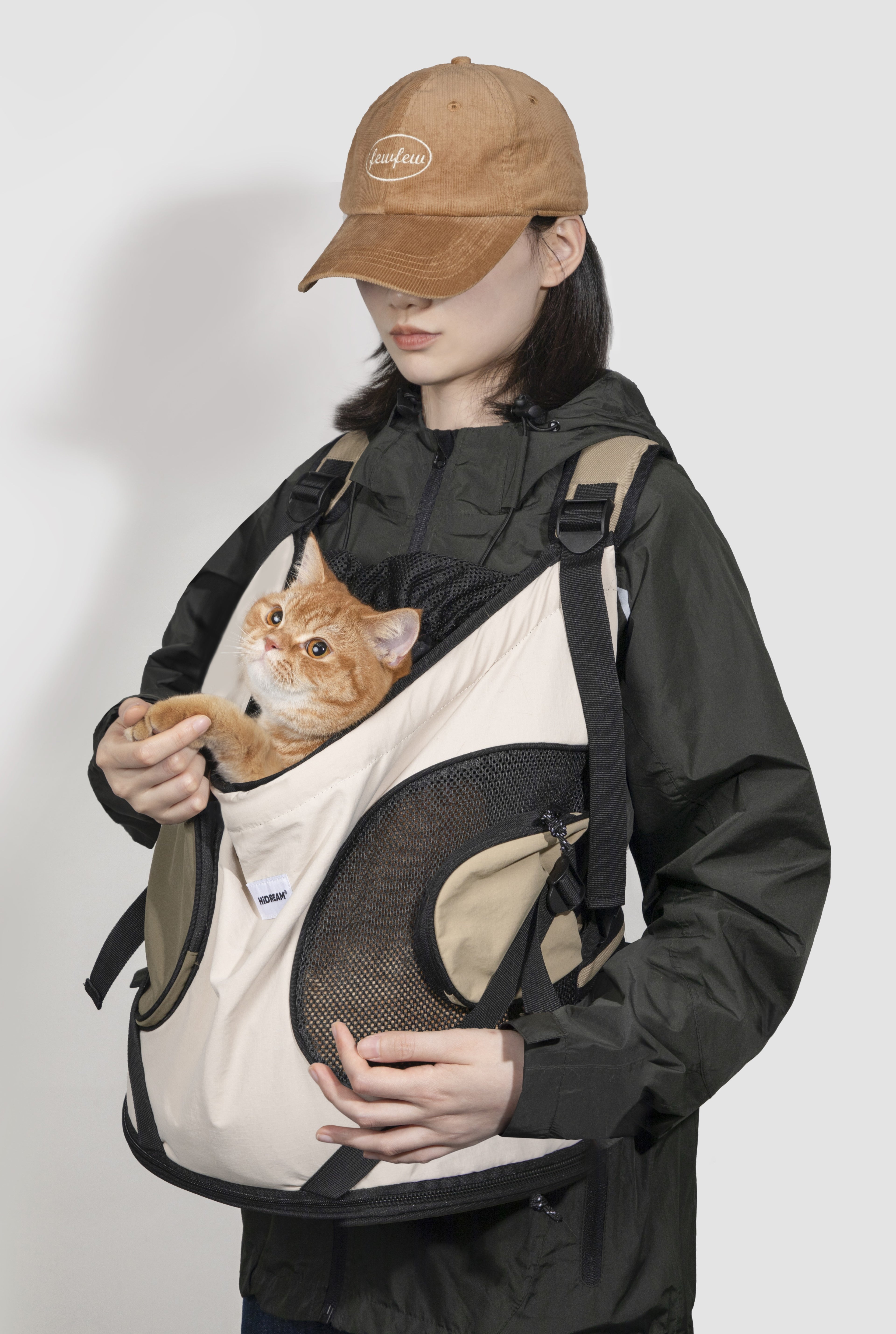 A person wearing a tan baseball cap and black jacket carries an orange tabby cat in a beige and black pet carrier backpack against a plain light gray background.