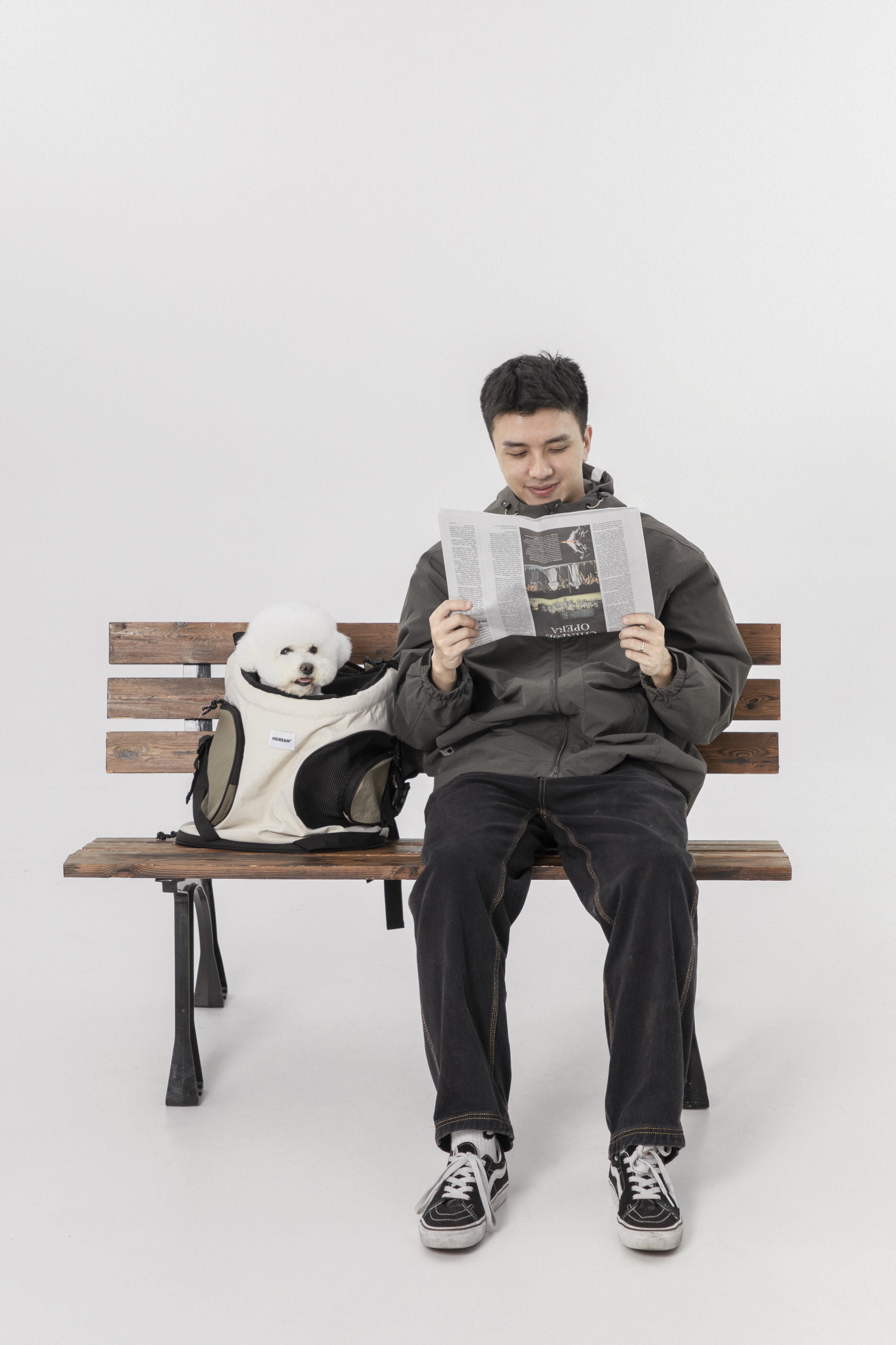 A man sits on a wooden bench reading a newspaper, with a small white dog in a pet carrier bag beside him. The man is wearing a dark gray hoodie, black pants, and black-and-white sneakers.