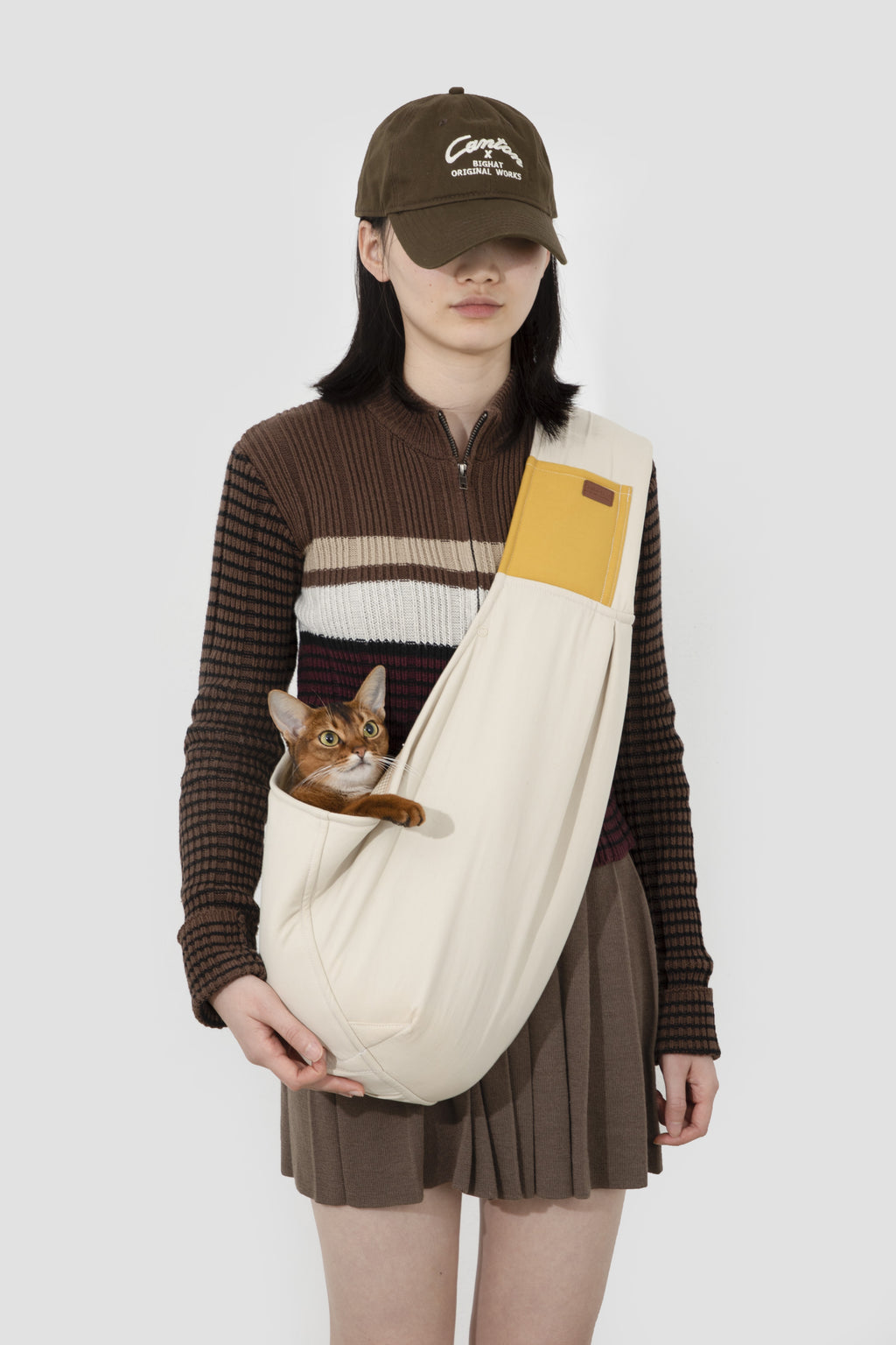 Person wearing a cap and holding a cat in a sling against a white background