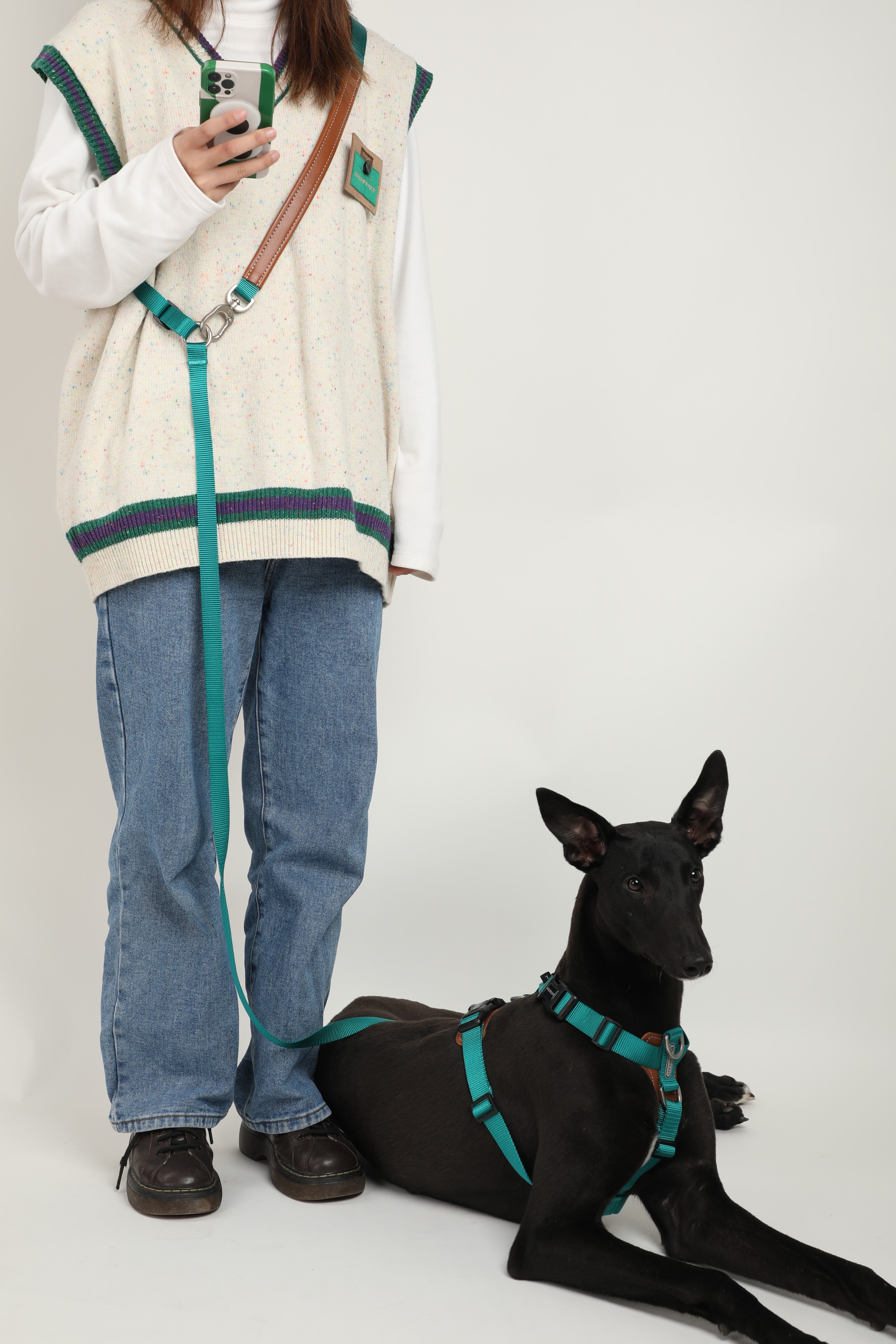 A person wearing a cream knit sweater vest with green trim, blue jeans, and brown shoes stands next to a black dog wearing a matching teal harness and leash, against a plain light gray studio background.