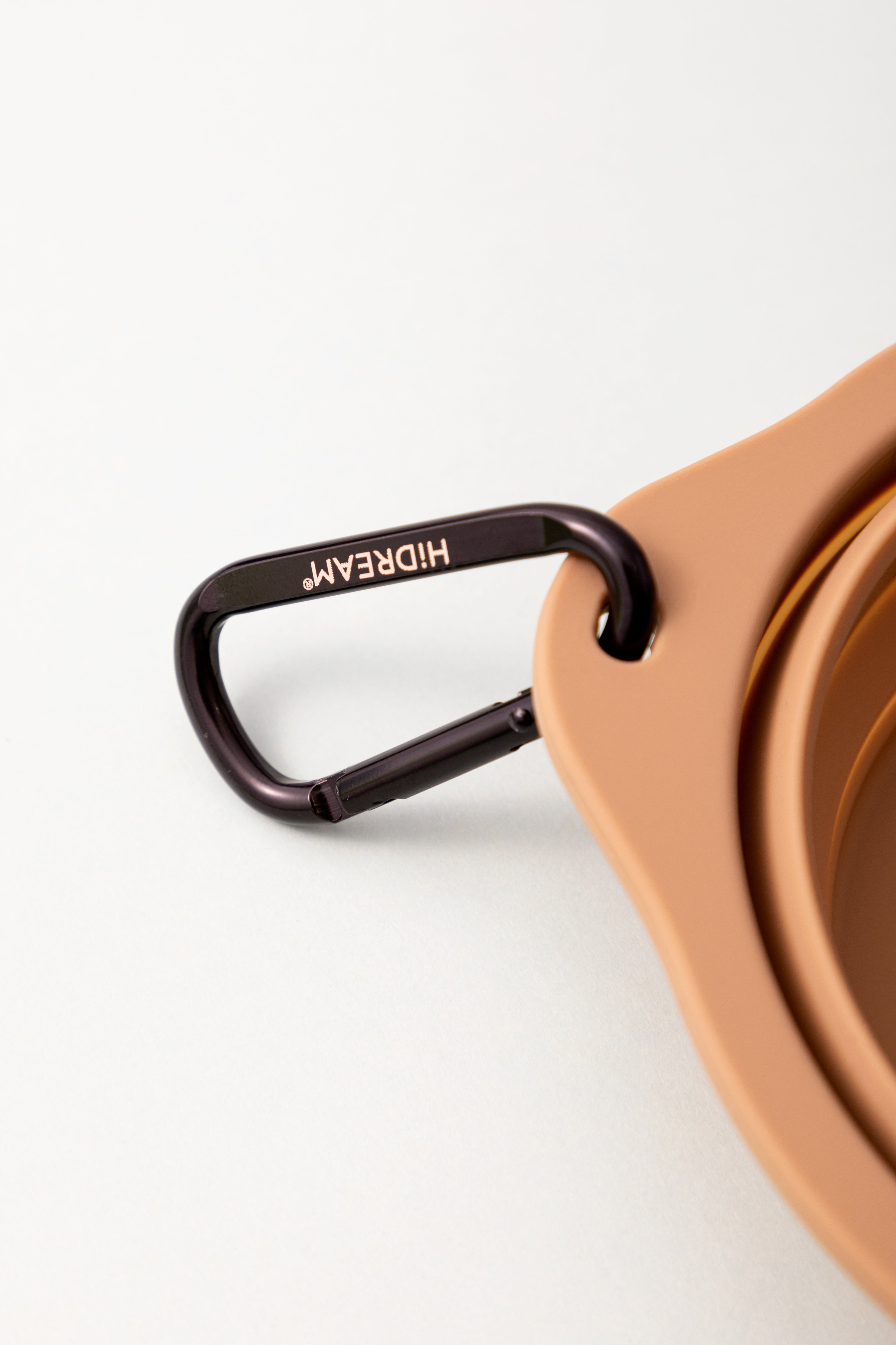 Close-up of a tan nested bowl or cup with a dark brown metal carabiner clipped to the rim. The carabiner is engraved with the brand name HIDREAM.
