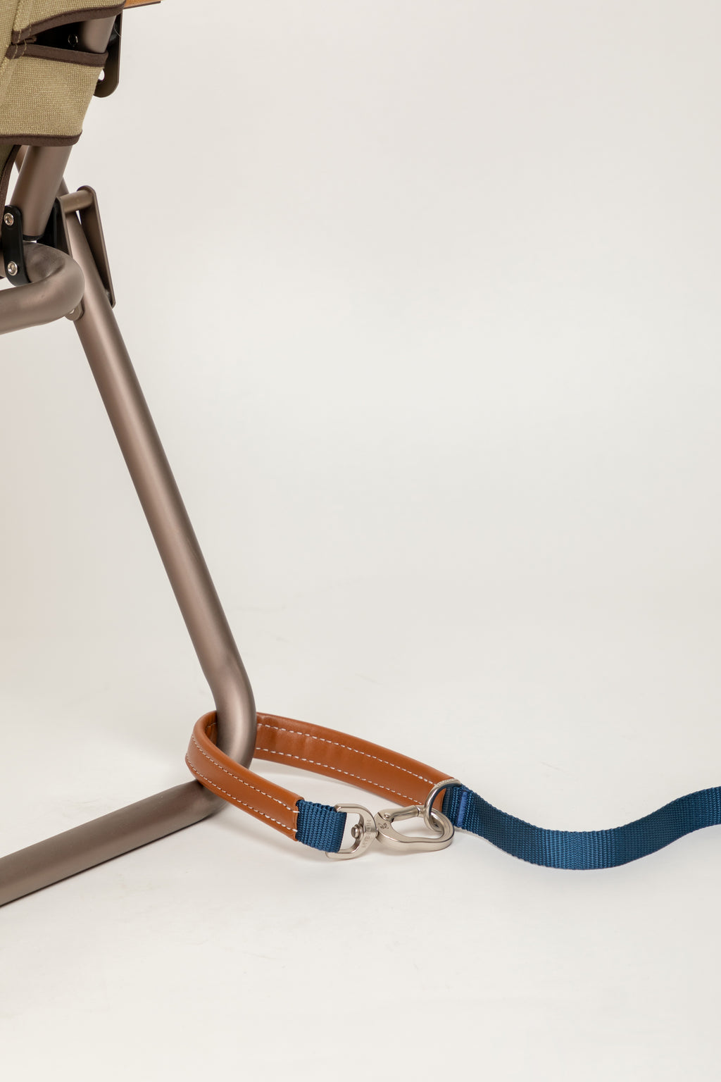 A close-up of a dog leash with a brown leather strap, silver metal hardware, and a blue webbed section, draped over the leg of a modern bronze chair against a clean white background.
