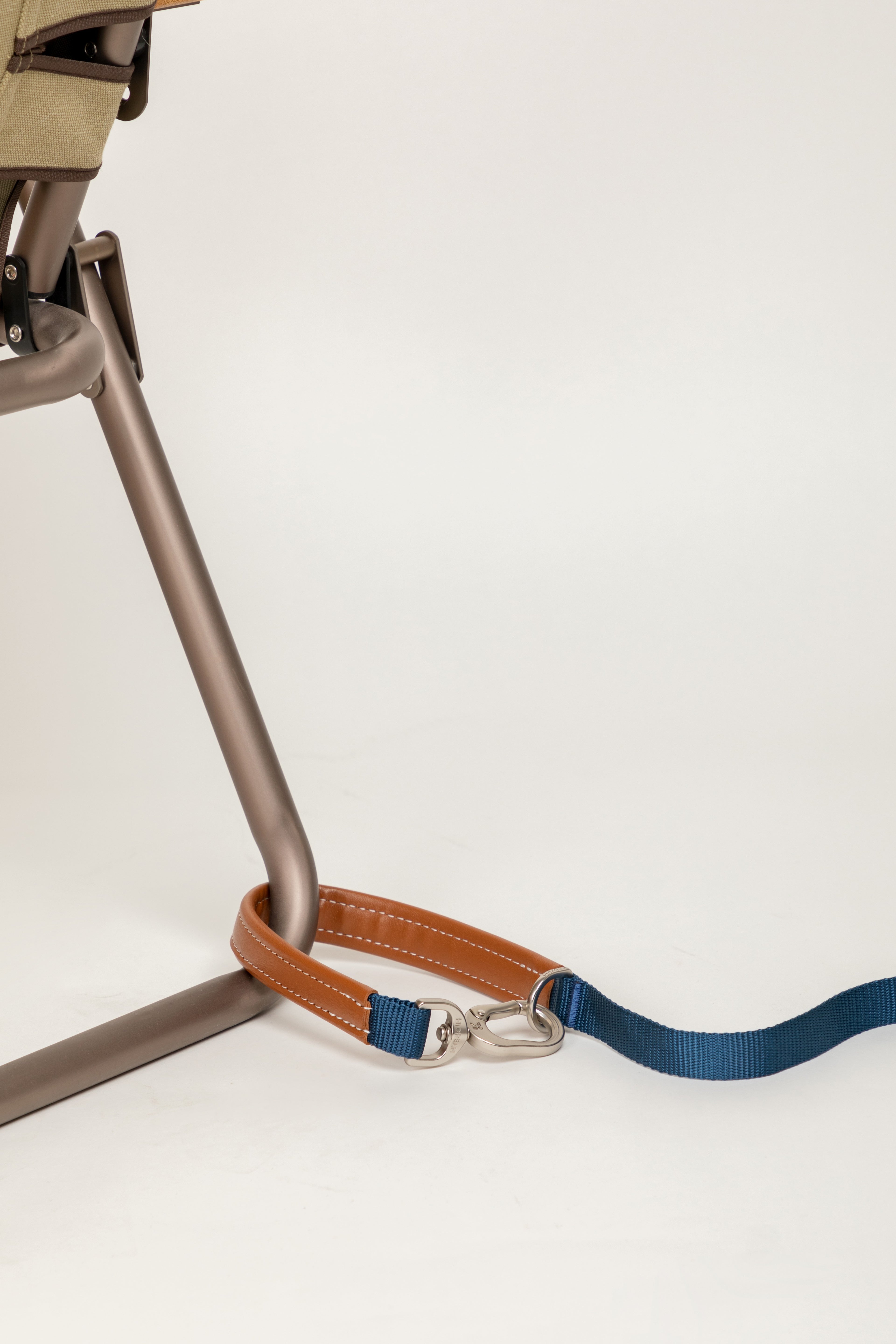 A close-up of a dog leash with a brown leather strap, silver metal hardware, and a blue webbed section, draped over the leg of a modern bronze chair against a clean white background.