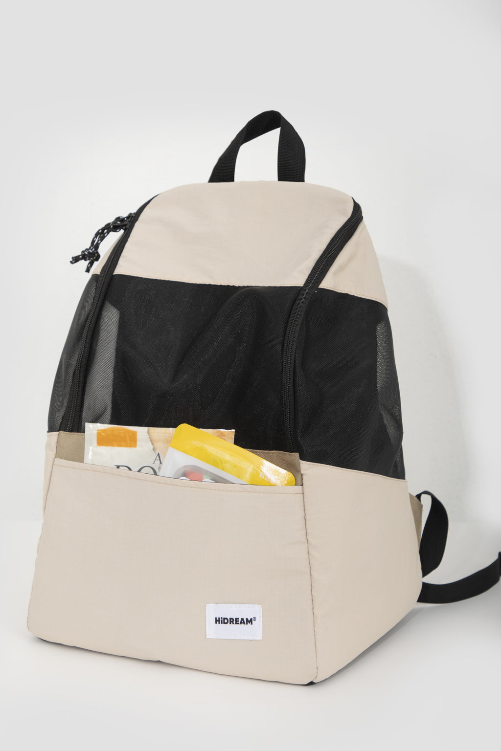 A colorblock backpack in beige and black, shown with its front pocket open to reveal some items inside. The brand name 'HIDREAM' is visible on a small white label on the lower front pocket.