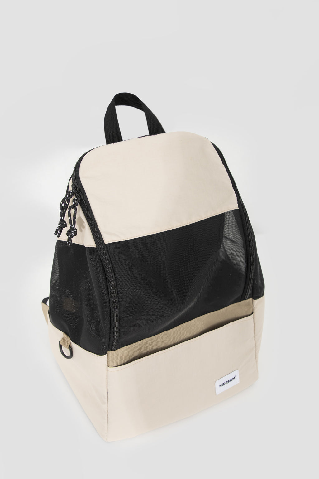 A color-blocked backpack in cream and black, featuring a top flap, a black mesh side pocket with a black tassel zipper pull, and a front slip pocket with a small white brand label.
