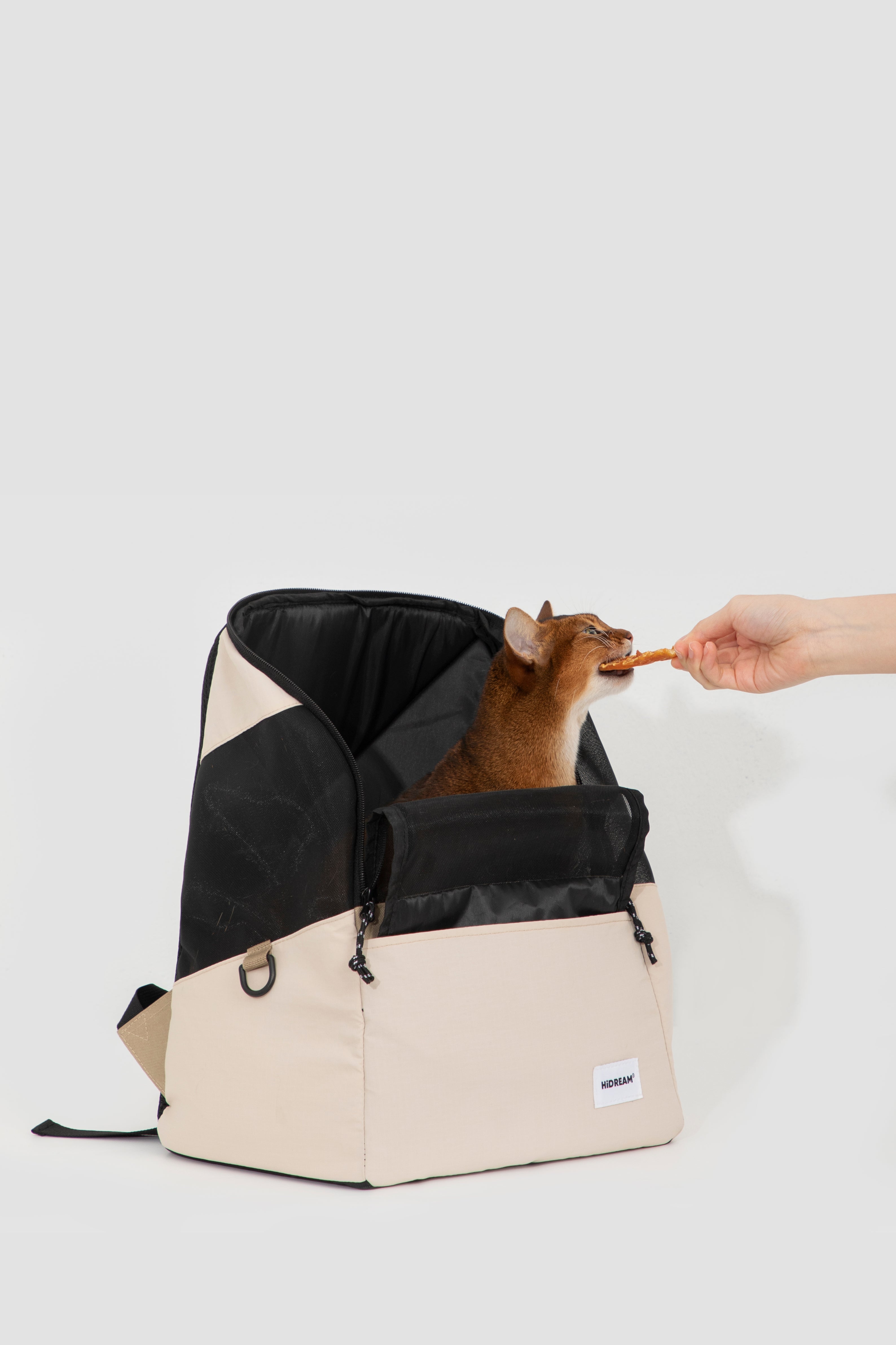 A beige and black pet carrier backpack with a small brown cat inside, while a human hand offers a treat to the cat, on a light gray background.